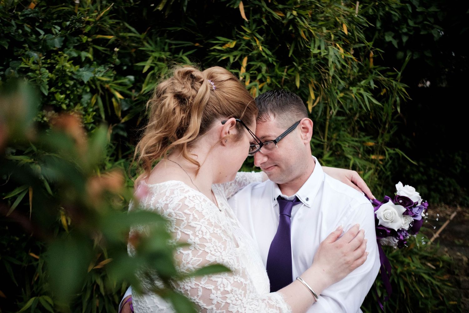 Bride and groom share a romantic forehead touch moment outdoors, surrounded by lush green foliage on their wedding day.