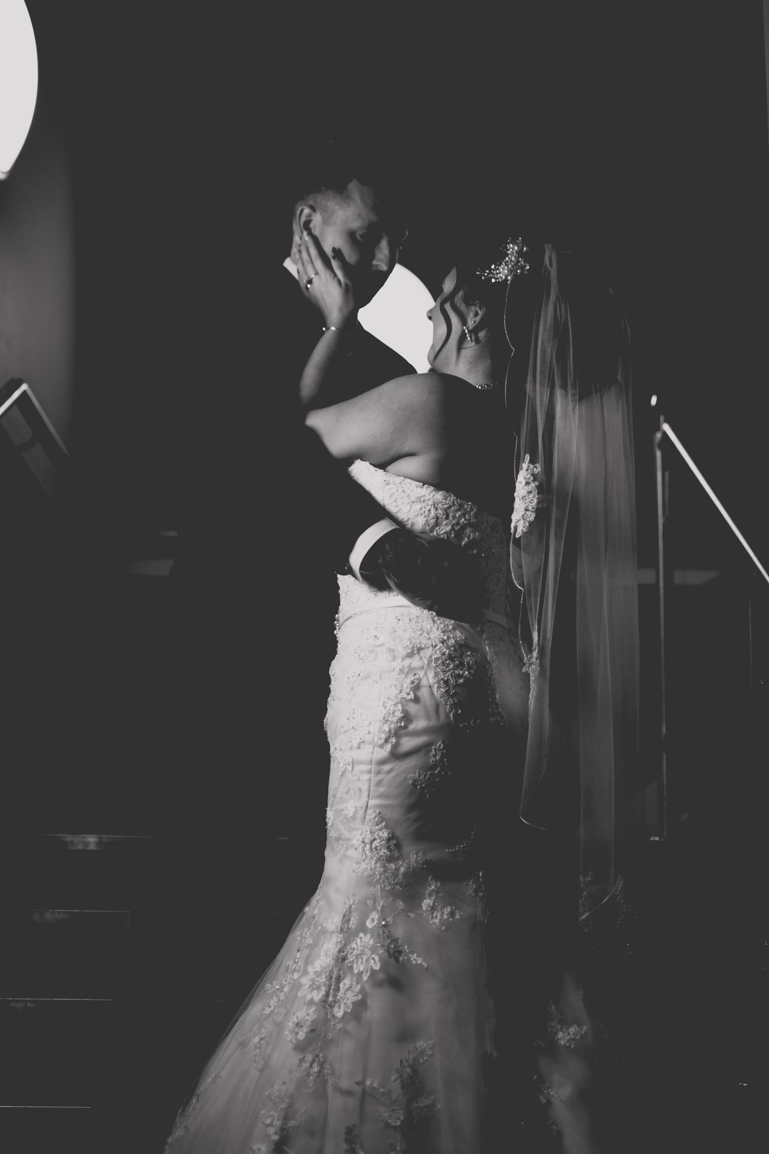 Black and white wedding photo of bride in lace gown and veil sharing romantic kiss with groom in dark setting.