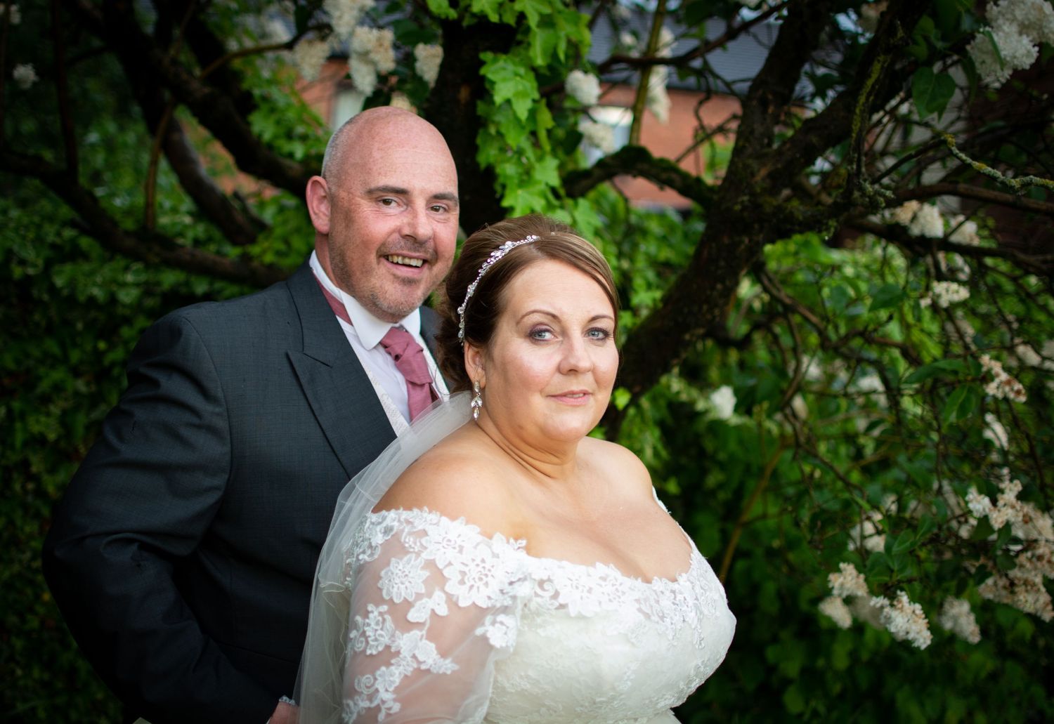 A bride and groom pose together outdoors under blooming spring trees, bride in lace gown, groom in dark suit.