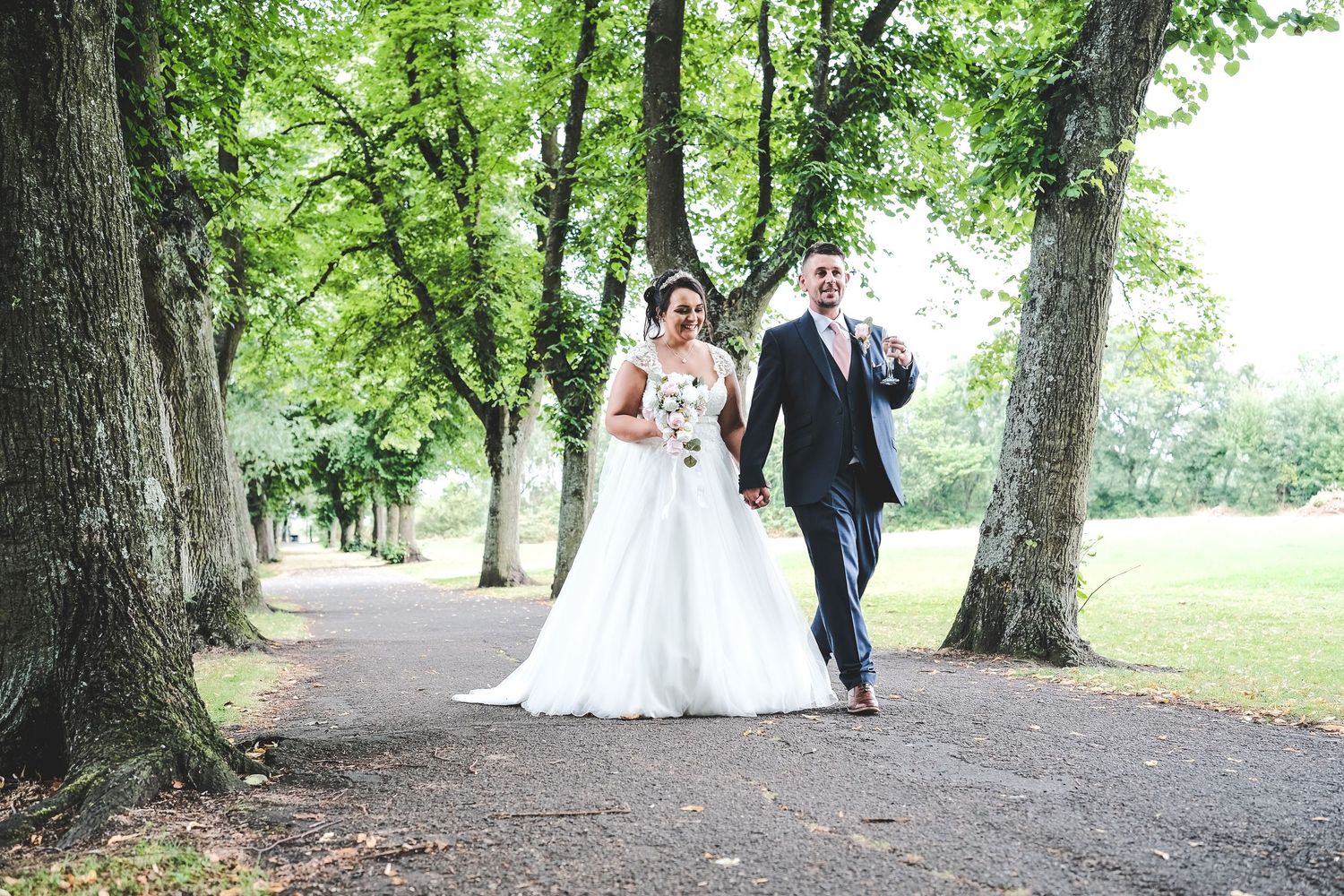 Bride and groom walking hand in hand along a tree-lined path on their wedding day.