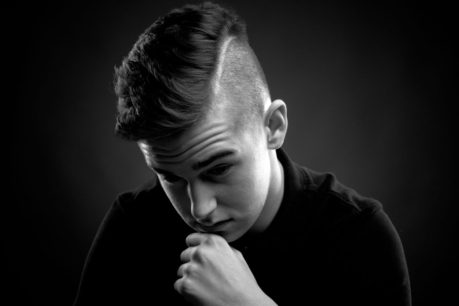 Black and white portrait of a young man in a black shirt, hand on chin, in a thoughtful pose against dark background.