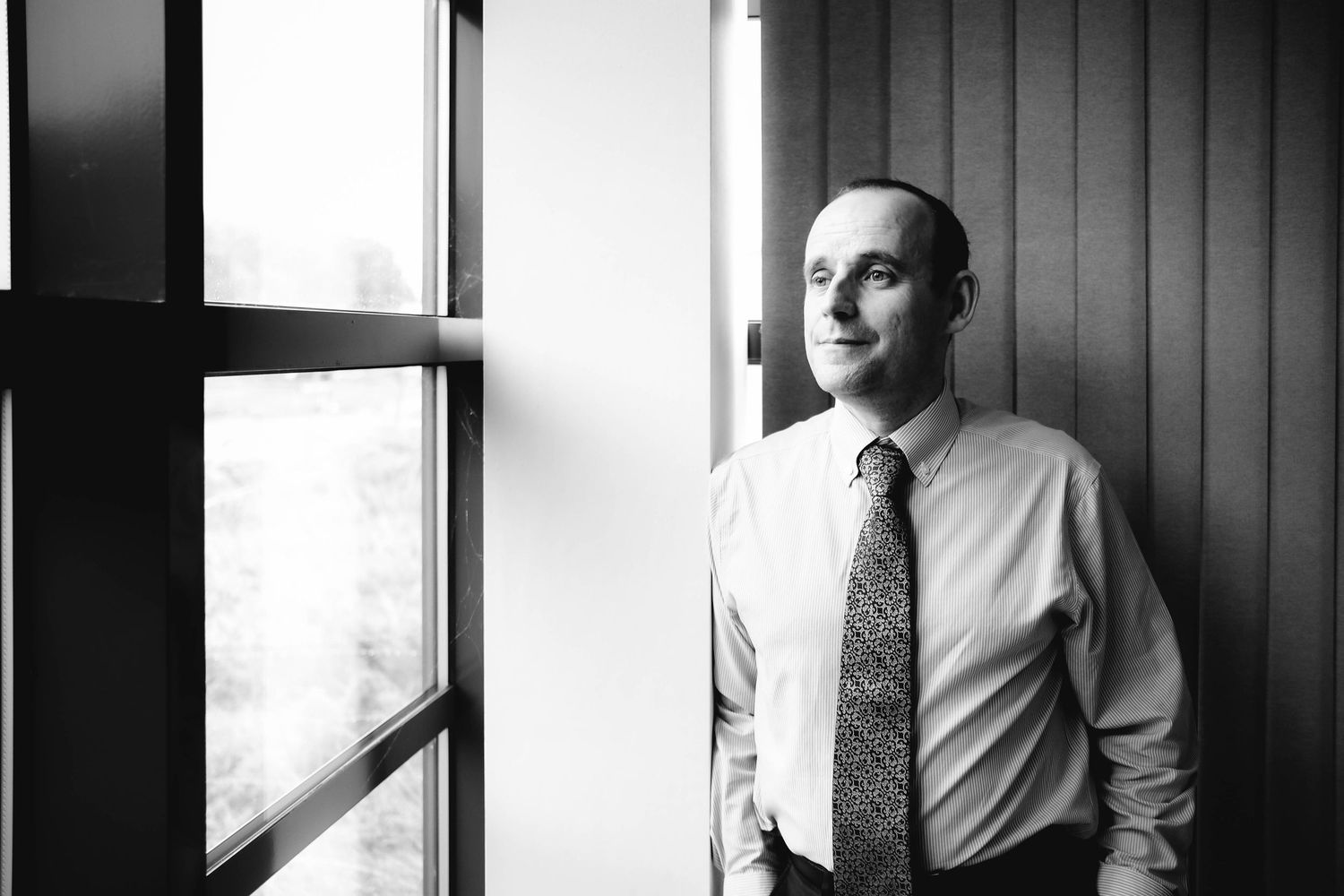 Black and white portrait of a businessman in shirt and tie standing by a window in an office building.