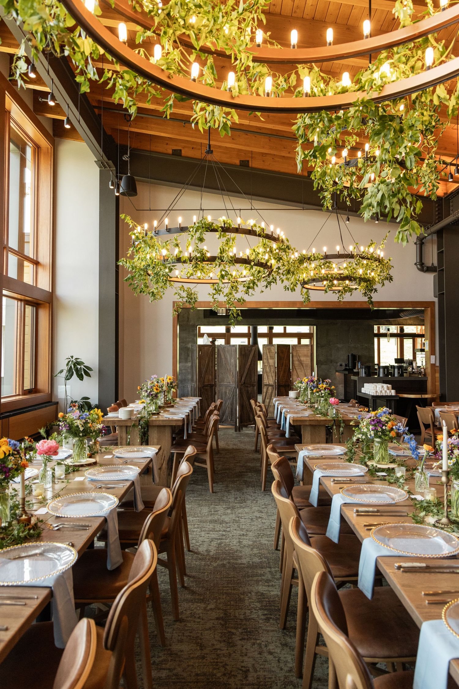 Elegant wedding reception venue with floral chandeliers, greenery ceiling decor, and long wooden tables set with colorful wildflowers.