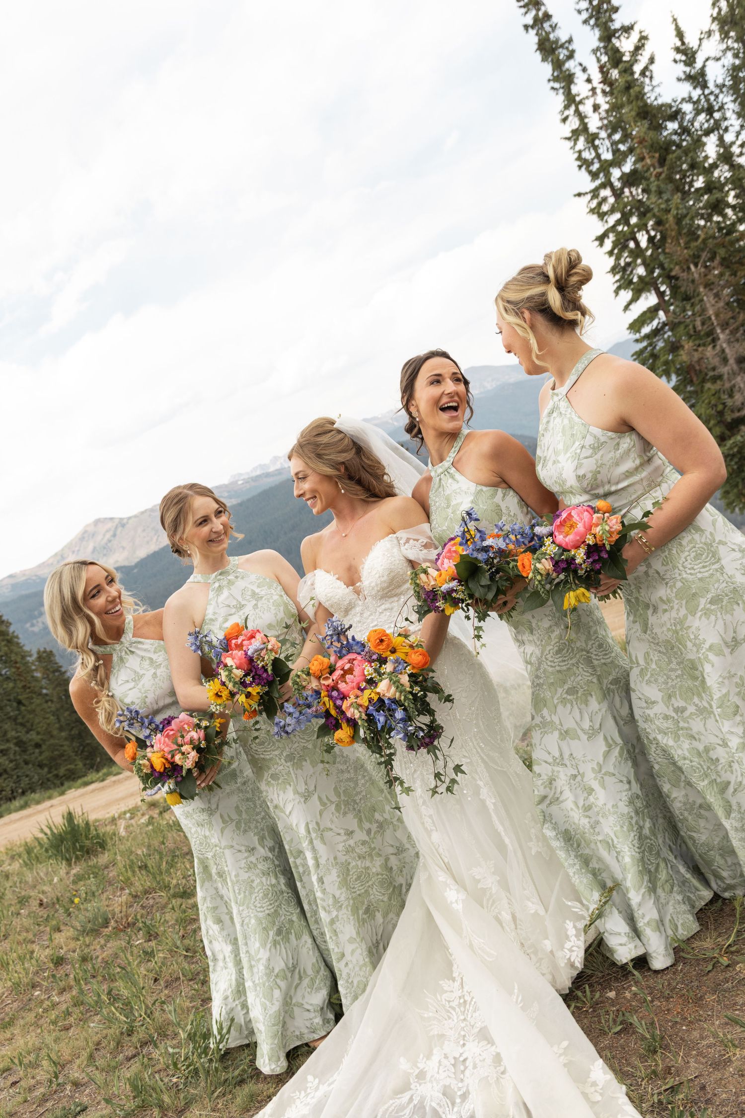 Bride and bridesmaids in sage green gowns laughing and holding colorful bouquets with mountain views behind them.