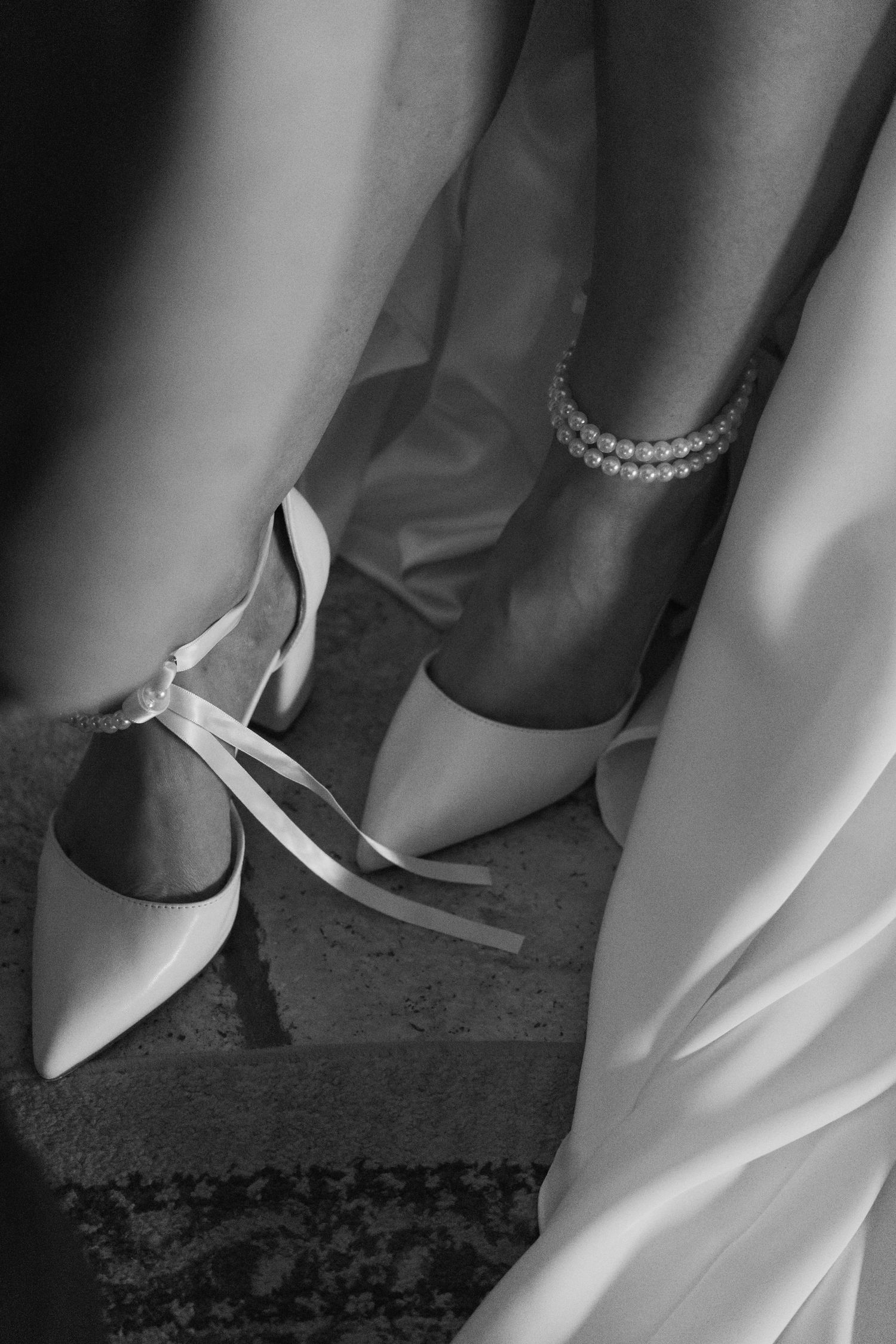 Black and white photo of a bride wearing elegant pointed-toe heels with ribbon ties and a pearl anklet bracelet.