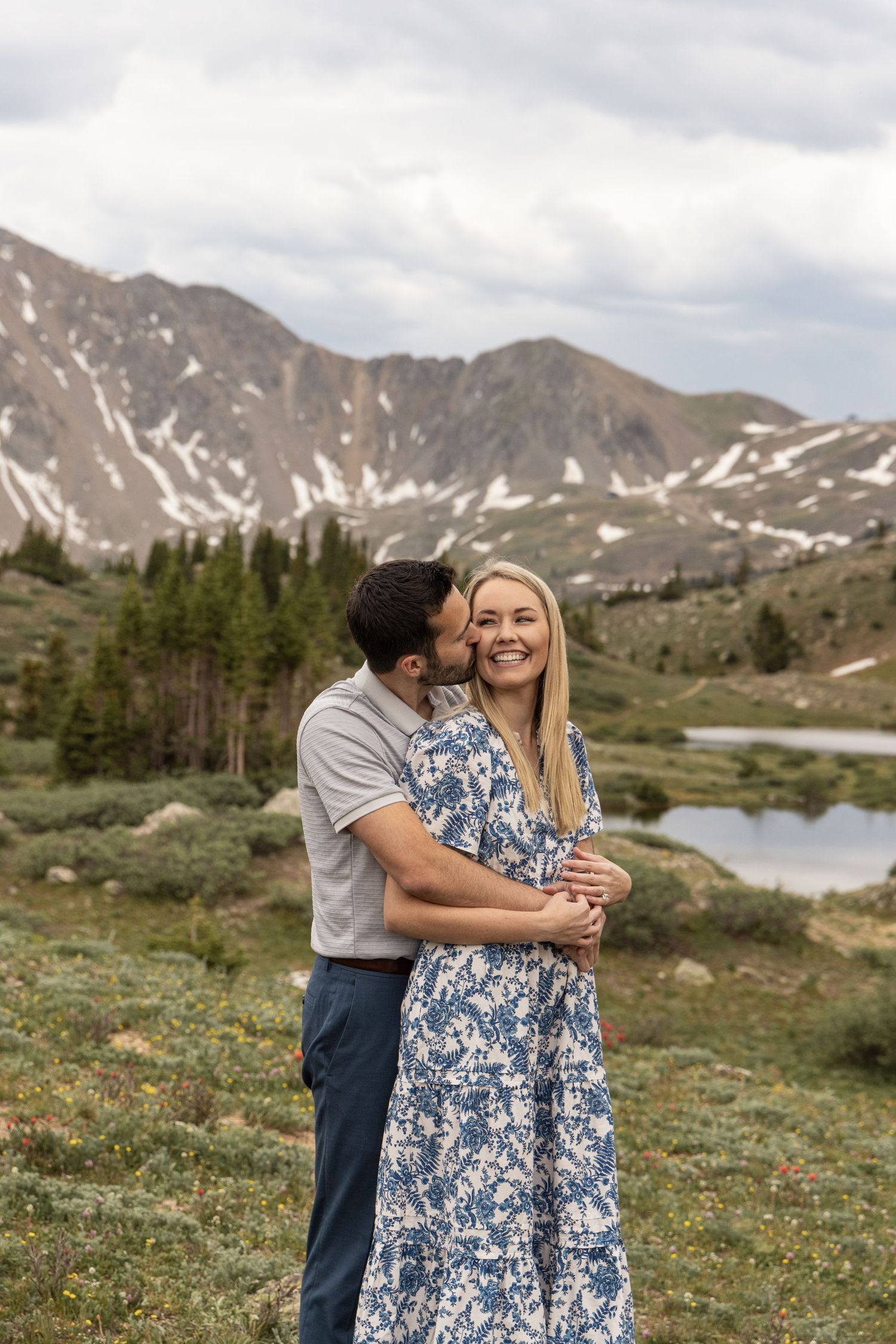A couple embraces against a stunning backdrop of snow-capped mountains and an alpine lake in a scenic mountain meadow.