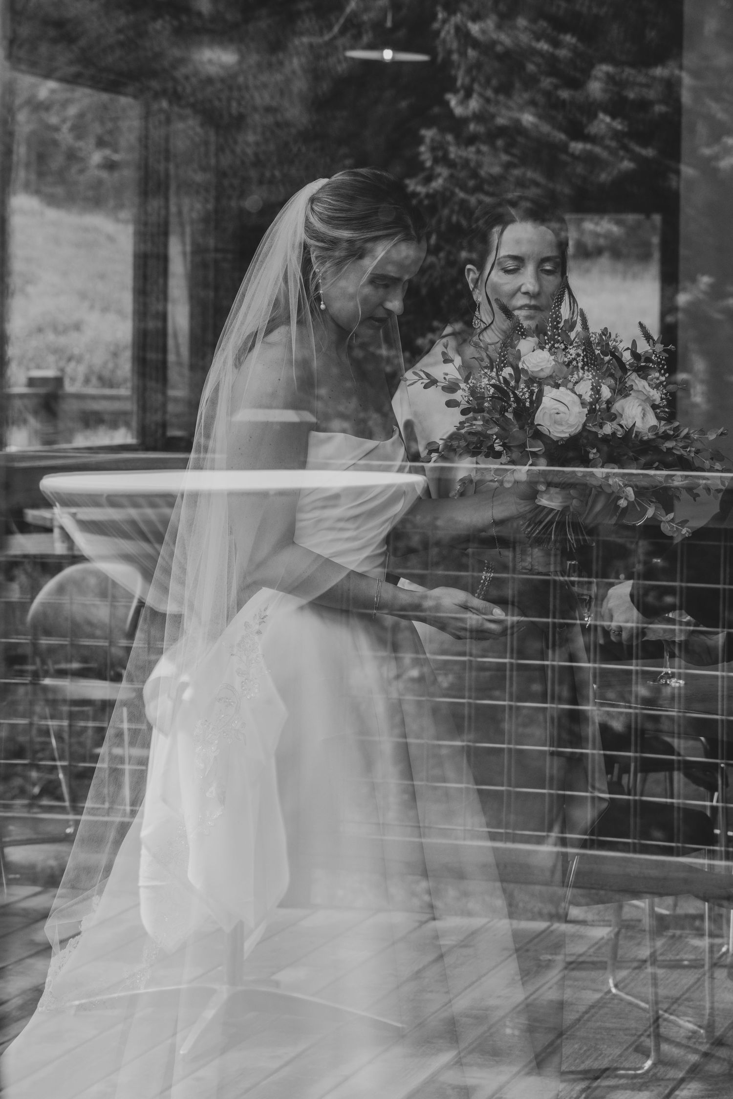 Black and white photo of two brides sharing a tender moment, one in a veil holding a bouquet, reflected in glass.