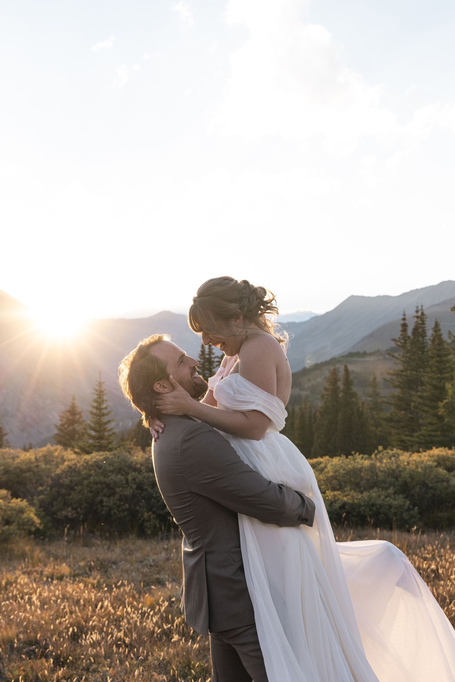 Newlywed couple embraces in a romantic sunset moment against a mountain landscape with evergreen trees.