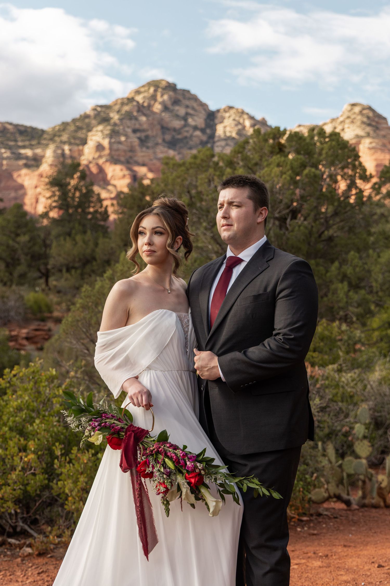 Bride and groom pose before red rock formations in Sedona, AZ, she holds a red floral bouquet.