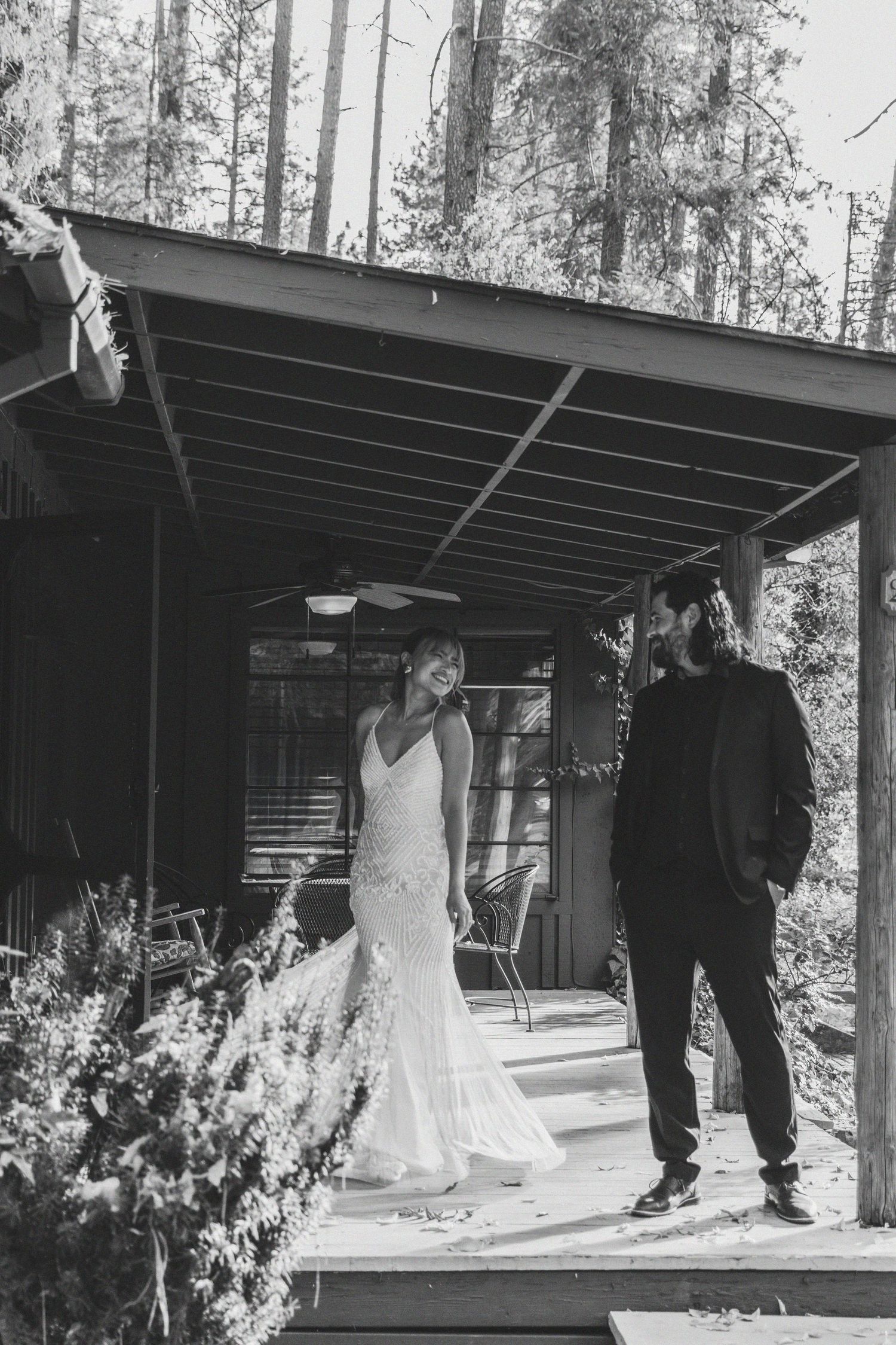 Black and white photo of a bride in a white gown and groom in dark suit on a rustic cabin porch surrounded by tall trees.