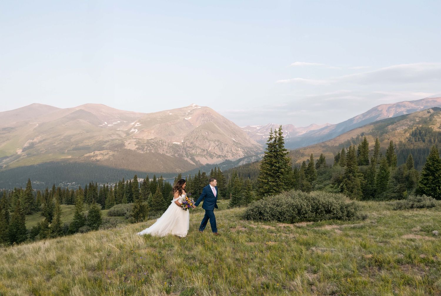 A couple enjoys a scenic mountain vista on a grassy meadow surrounded by evergreen trees and rolling peaks in Colorado.