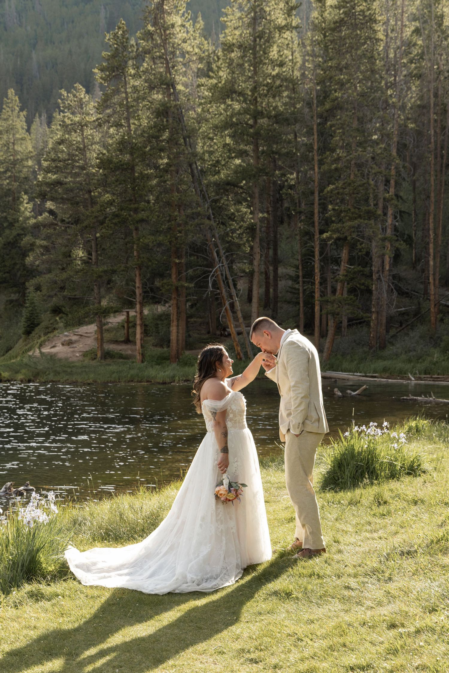 Bride and groom share a romantic moment by a mountain lake, surrounded by tall pine trees in a lush forest setting.