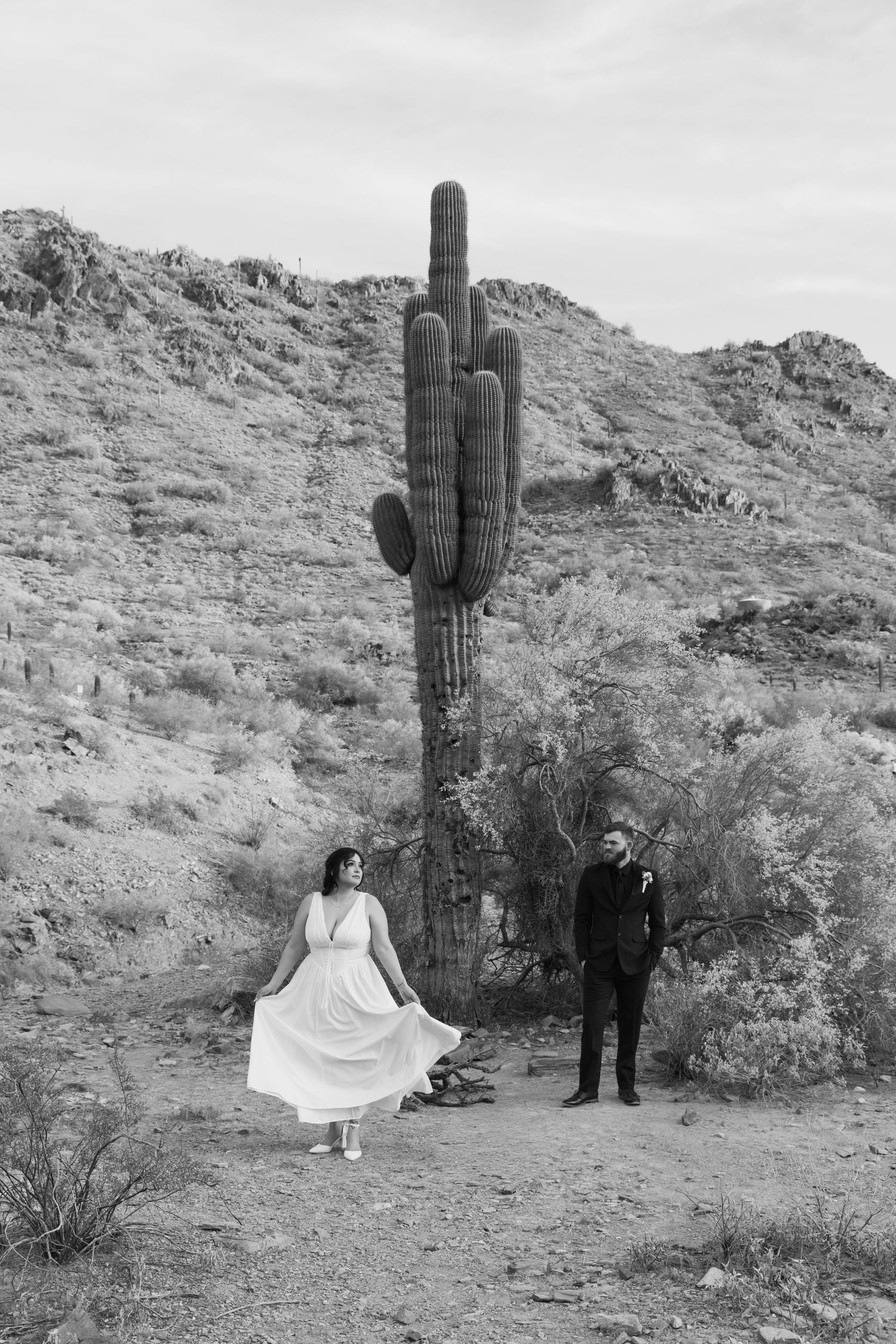 Black and white desert elopement photo of bride in white dress and groom near tall saguaro cactus on rocky hillside.