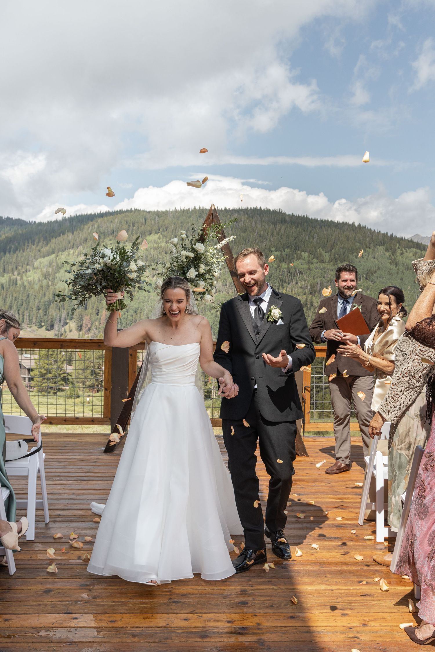 Newlywed couple walks down outdoor wedding aisle on mountain deck as guests toss flower petals in celebration.