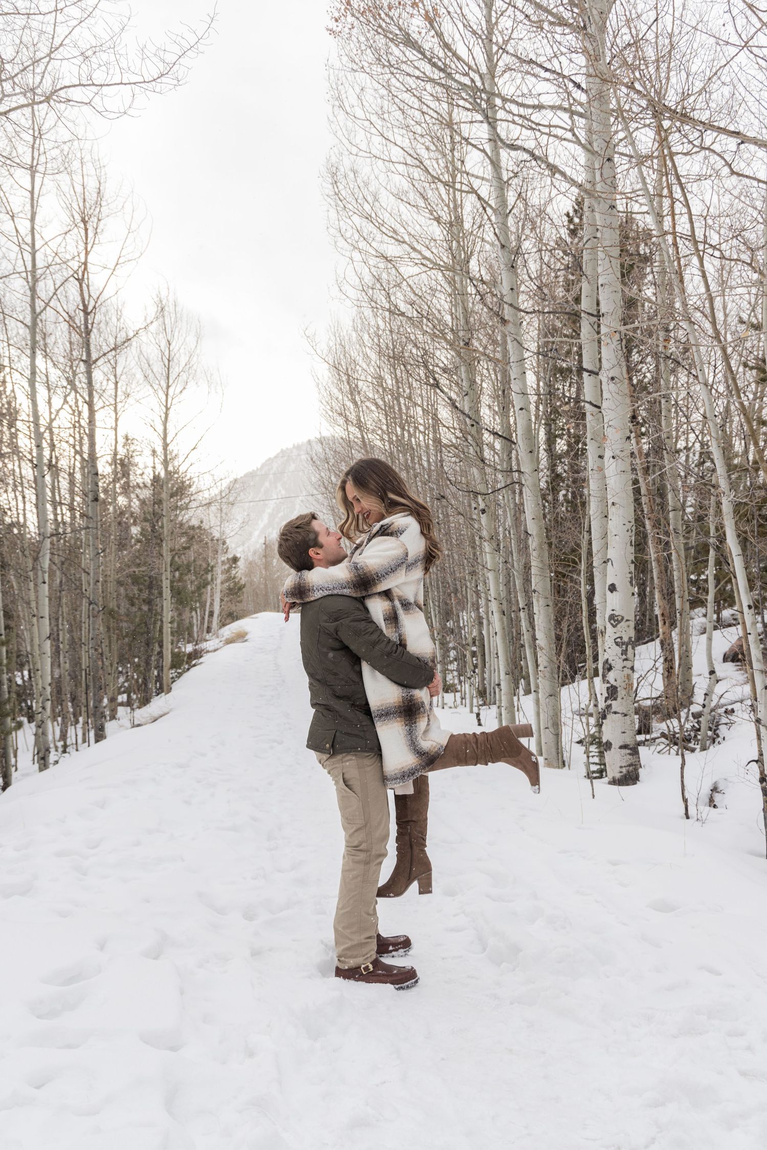 A couple embraces in a snowy winter forest with bare aspen trees and a mountain view in the background.