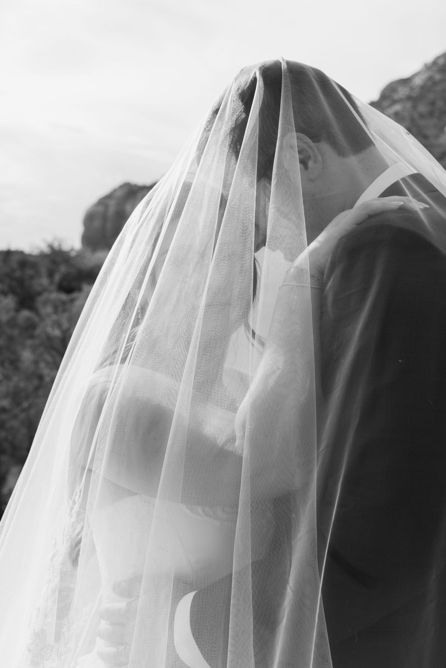 Black and white photo of bride and groom sharing a kiss under a flowing wedding veil outdoors.