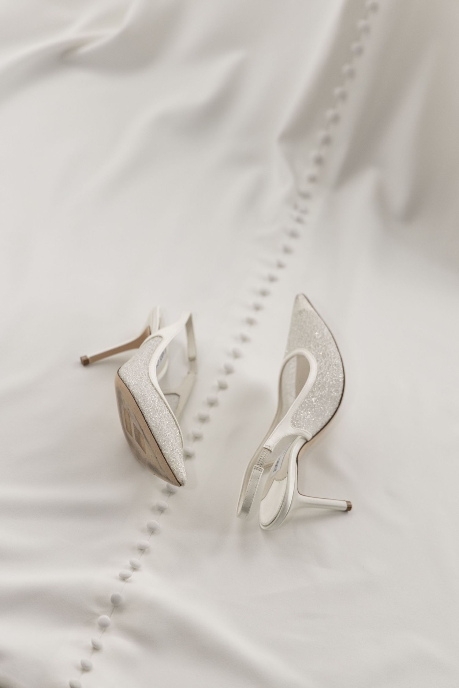 Elegant white bridal high heel shoes with lace detail resting on a white wedding dress with pearl buttons.