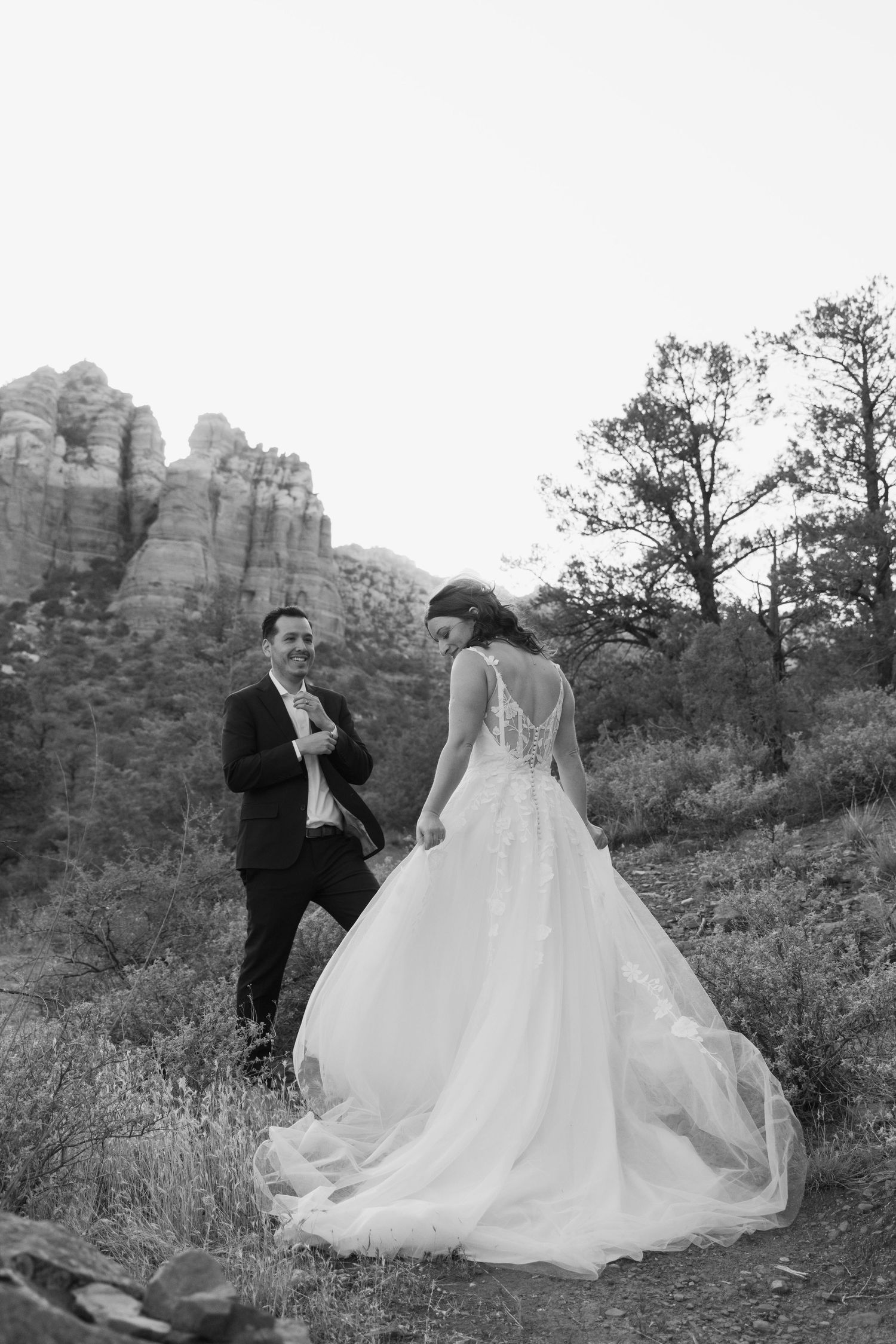 Black and white photo of bride and groom walking among red rocks in Sedona, Arizona, bride in flowing ballgown.