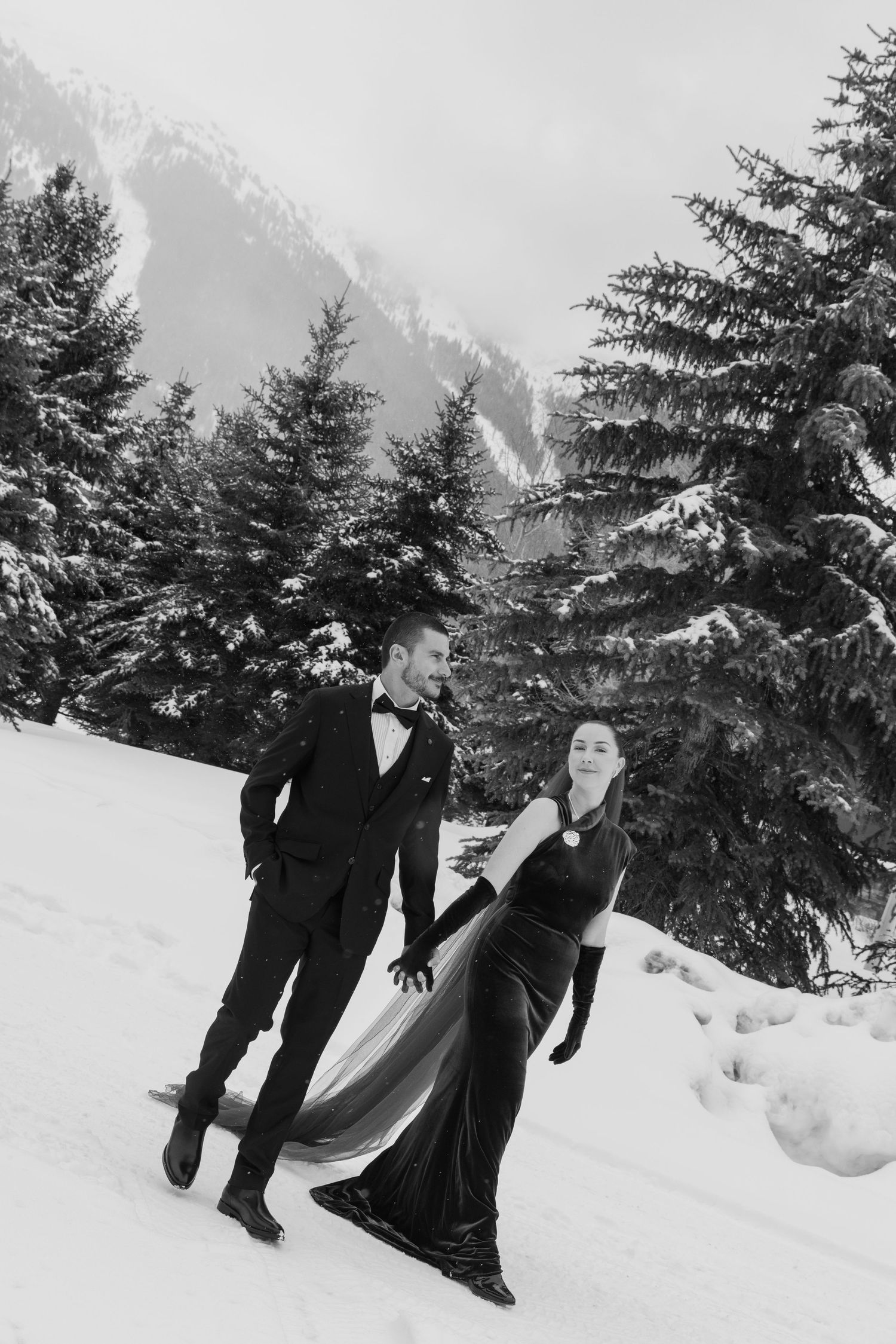 Elegant couple in black formal wear walking through snowy mountain landscape with snow-covered pine trees.
