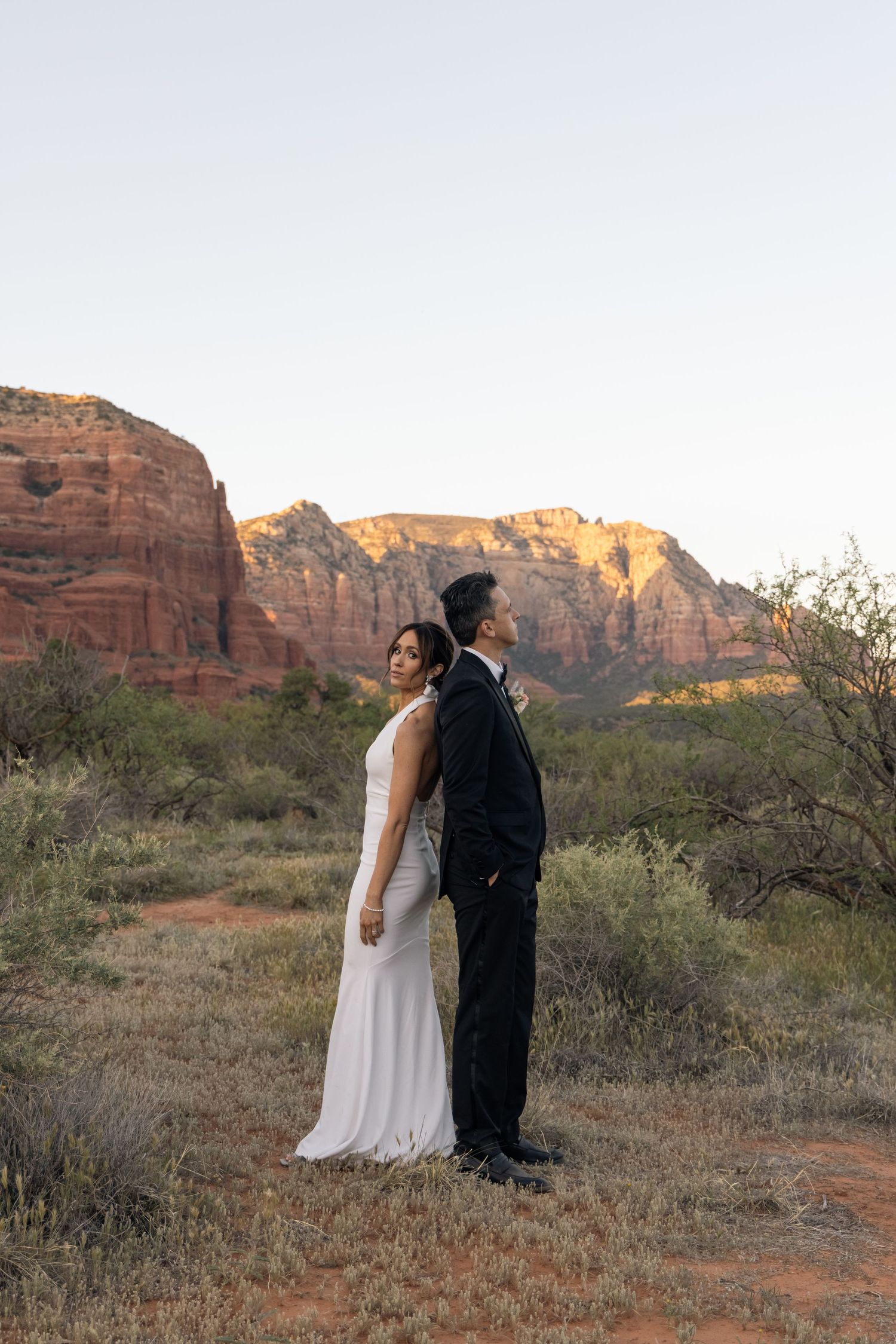 A couple in wedding attire stands together against majestic red rock formations in the desert at sunset.