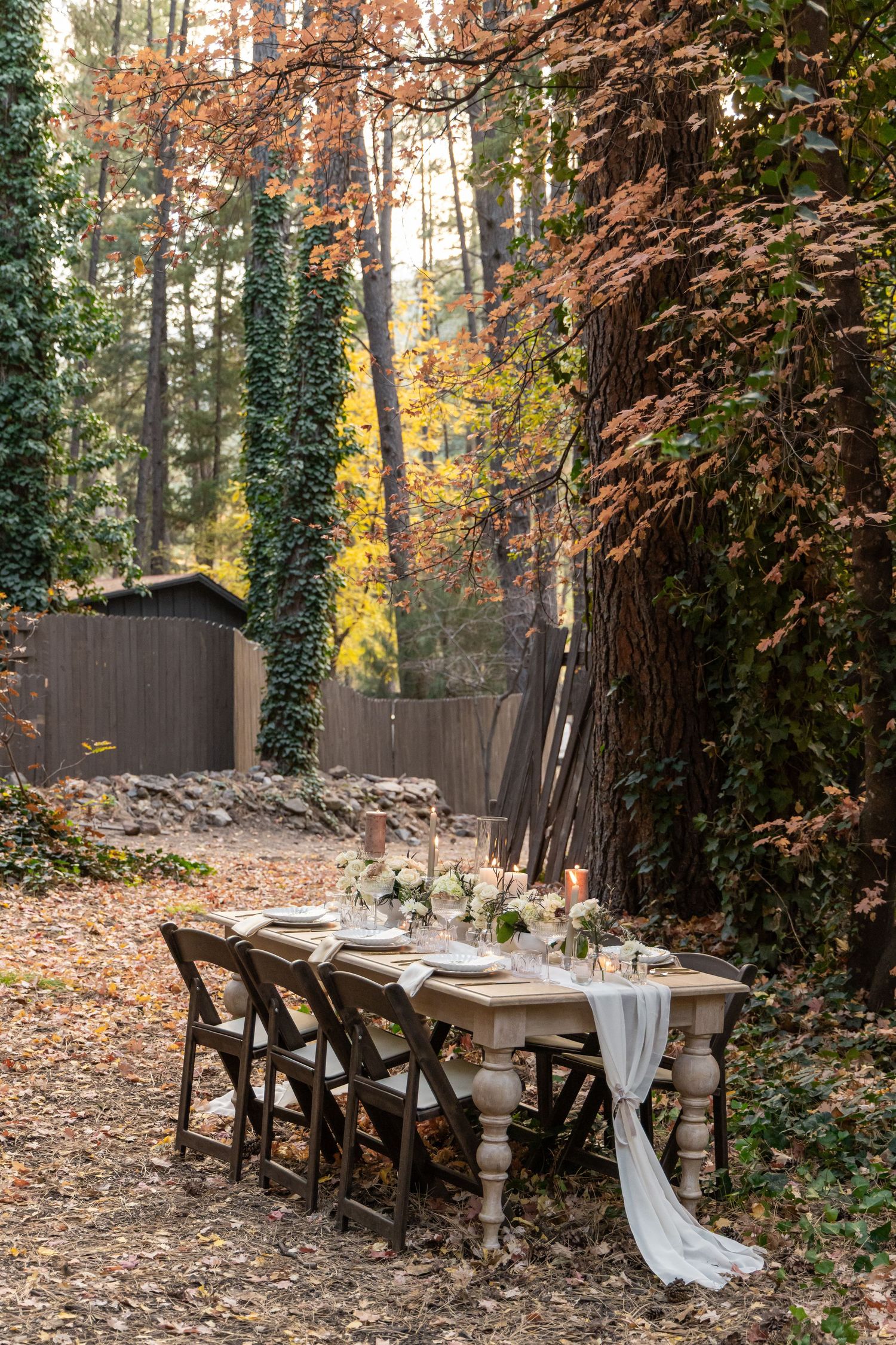 Rustic outdoor dining table with white linens and wooden chairs set in an autumn forest with fallen leaves and brown foliage.