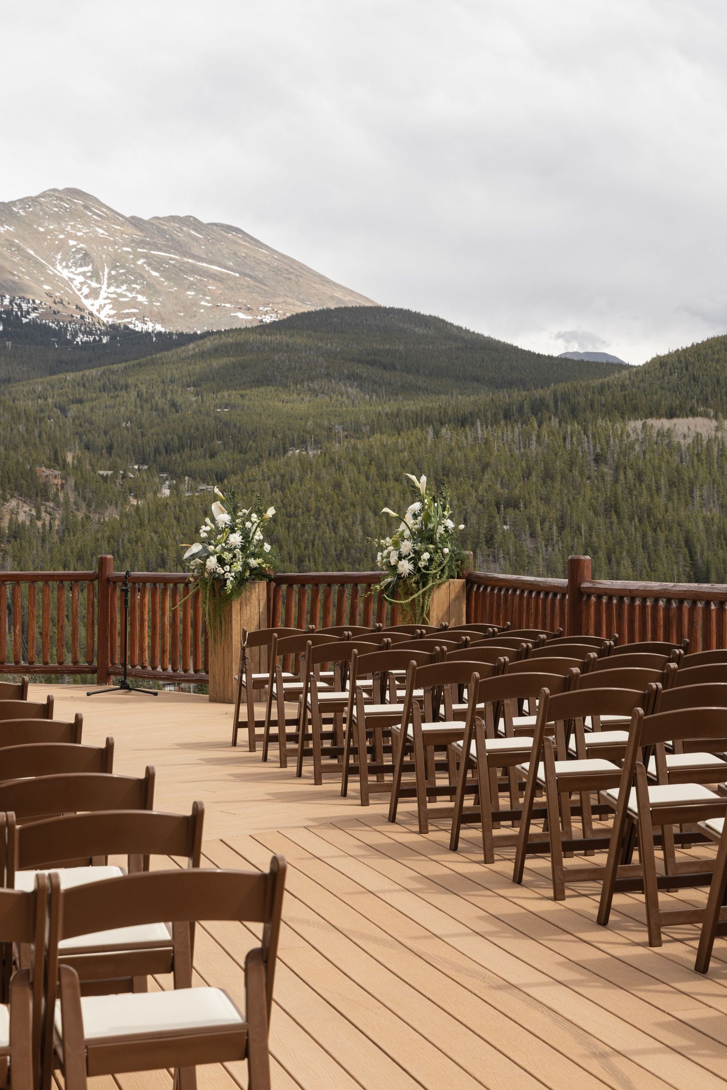 Outdoor wedding ceremony setup on wooden deck with brown folding chairs, floral arrangements, and scenic mountain forest backdrop.