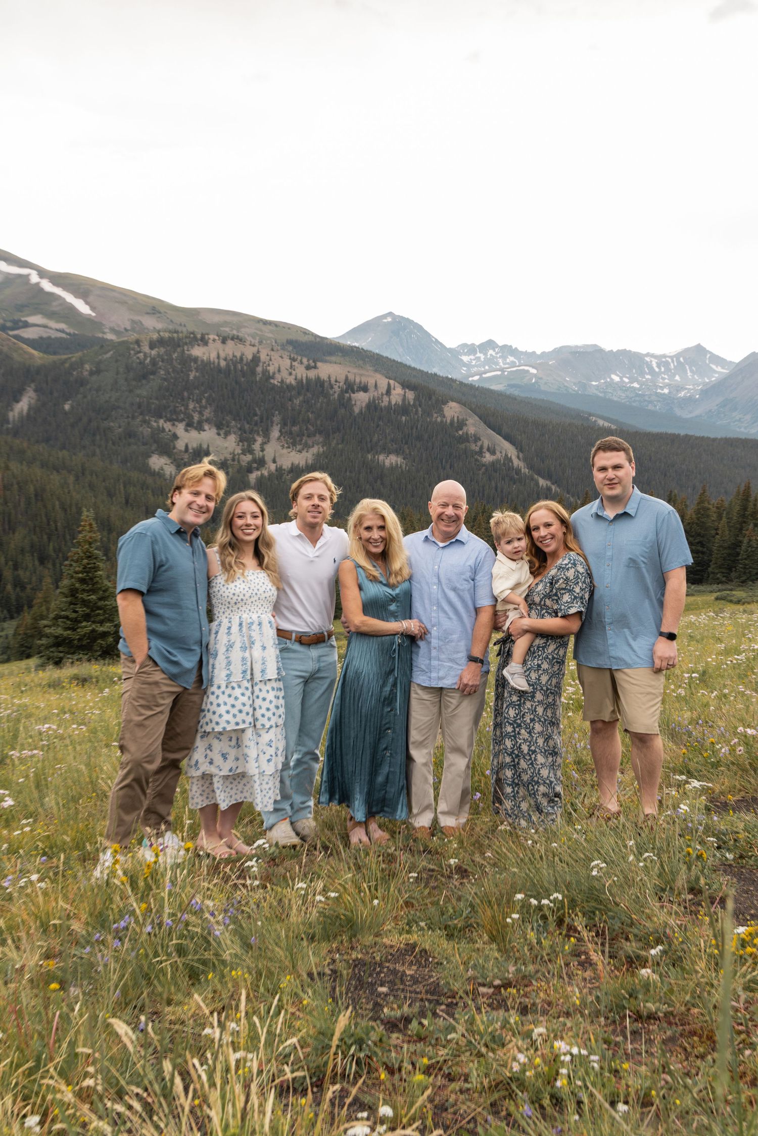 A group stands together on a grassy hillside with majestic snow-capped mountains and evergreen forests in the background.