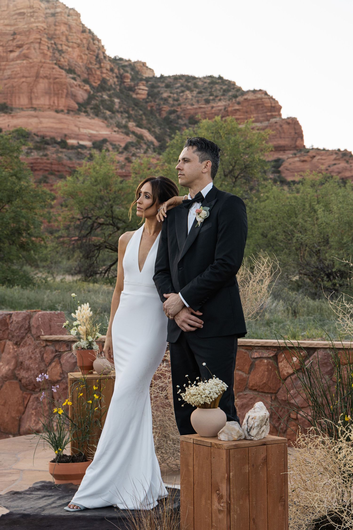 Couple in wedding attire posing against Sedona red rock backdrop, bride in white gown, groom in black tuxedo.
