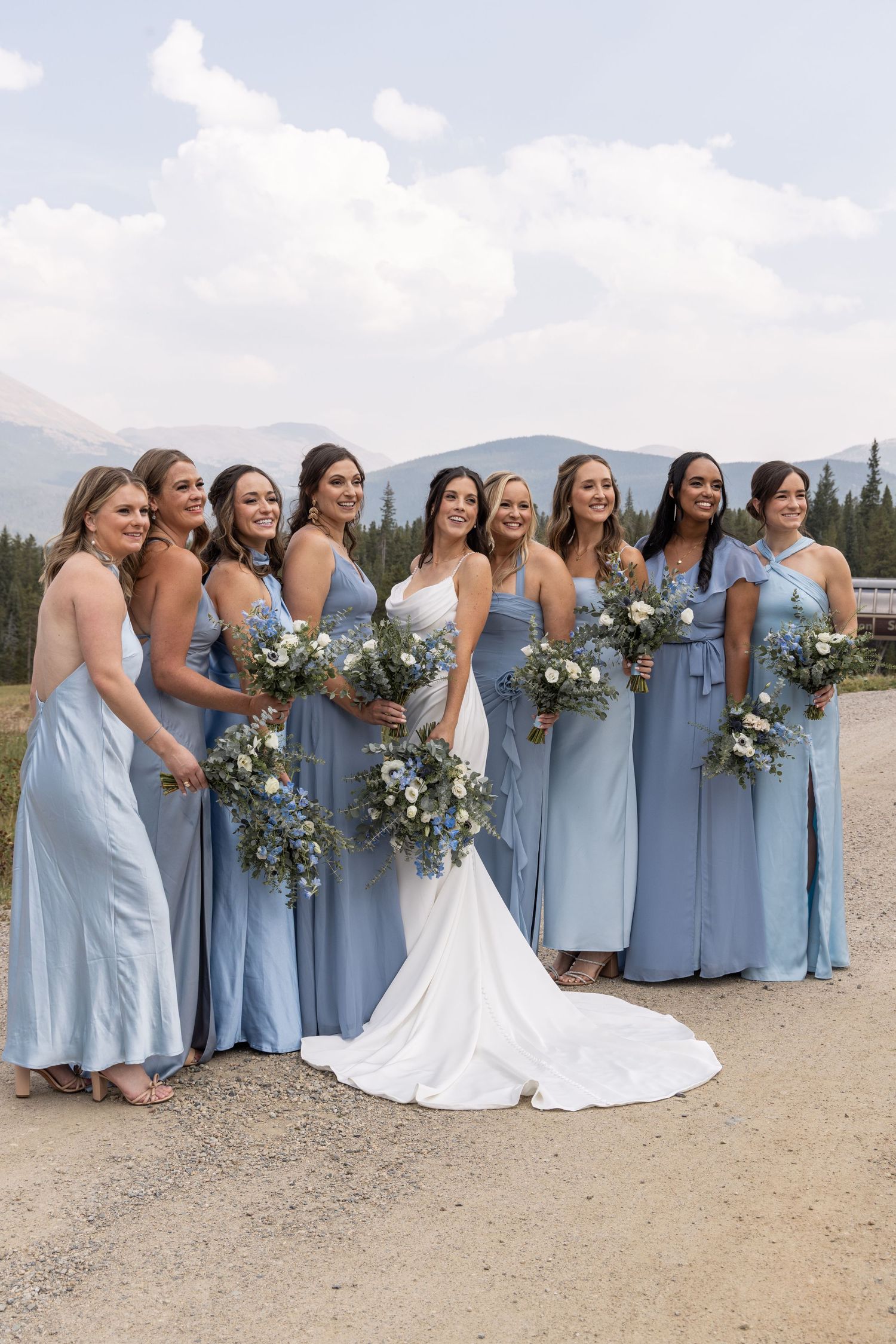 Bridesmaids in light blue dresses holding white and blue floral bouquets pose with bride in white gown on mountain path.