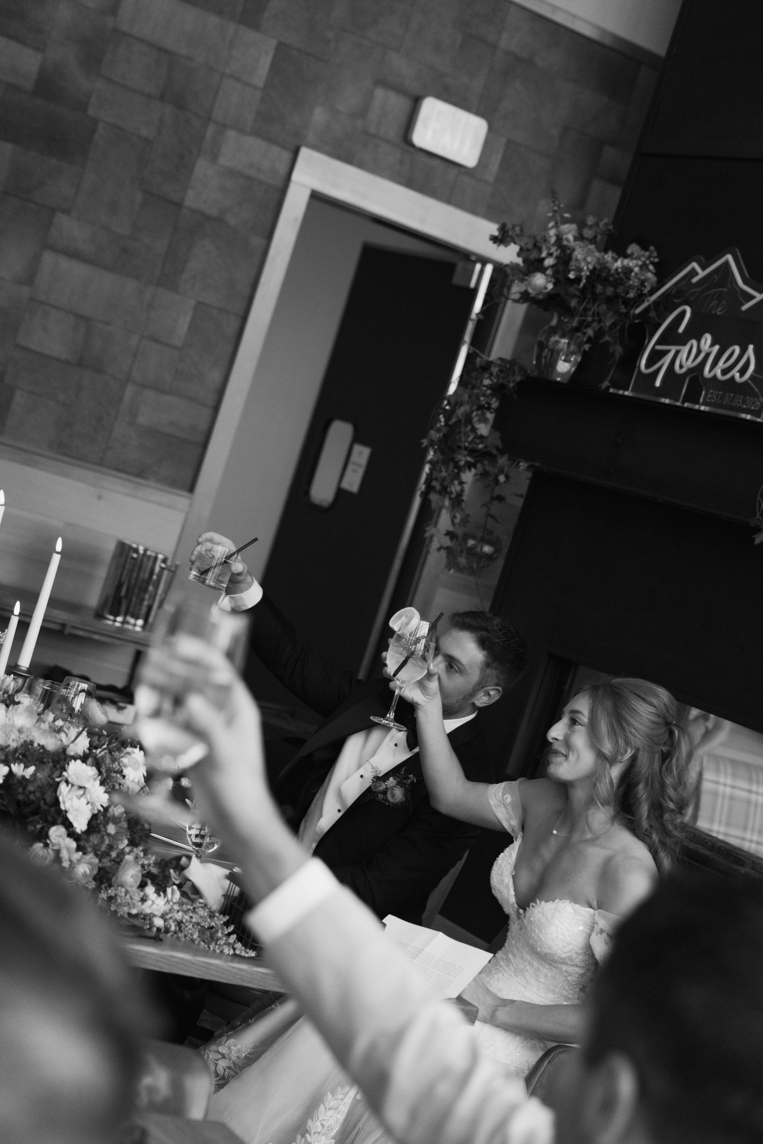 Black and white wedding reception photo of smiling bride and groom toasting with guests at elegantly decorated table.