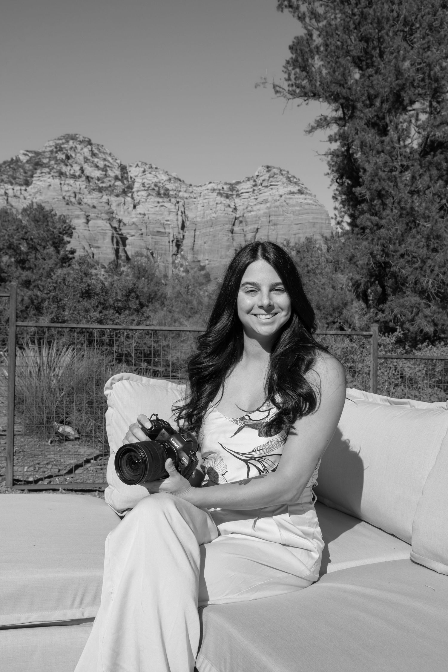 A person in a white dress sits outdoors on a sunny day with mountains and trees visible in the background.