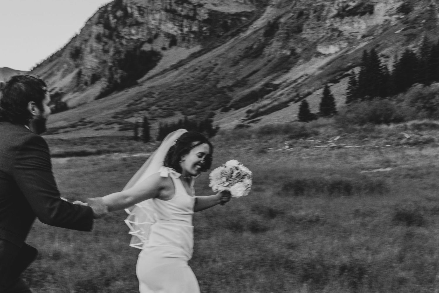 A couple enjoys a playful moment in a mountain meadow with rugged peaks rising in the background.