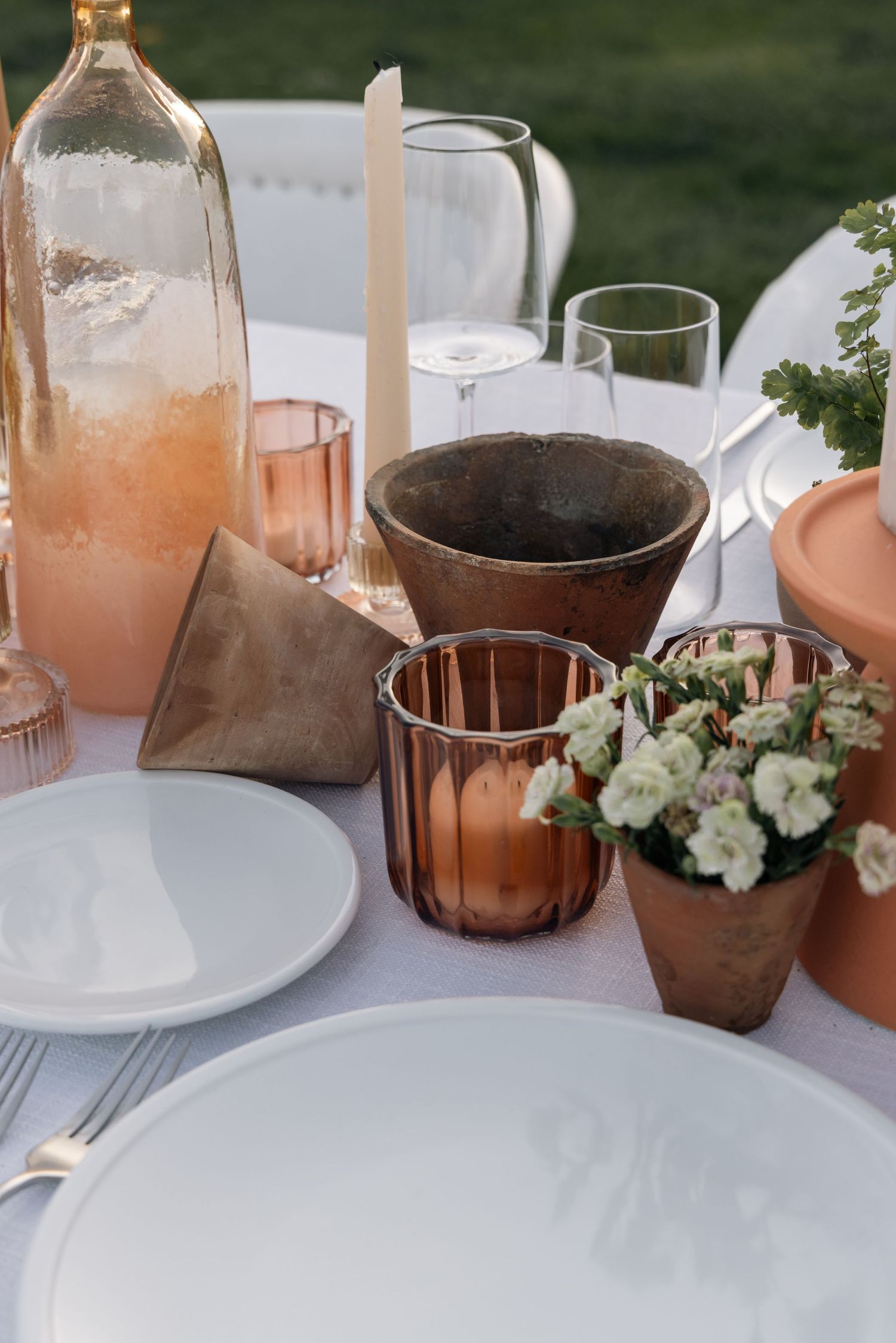 Elegant outdoor table setting with terracotta pots, white flowers, candles, and rose-tinted glassware on a white tablecloth.