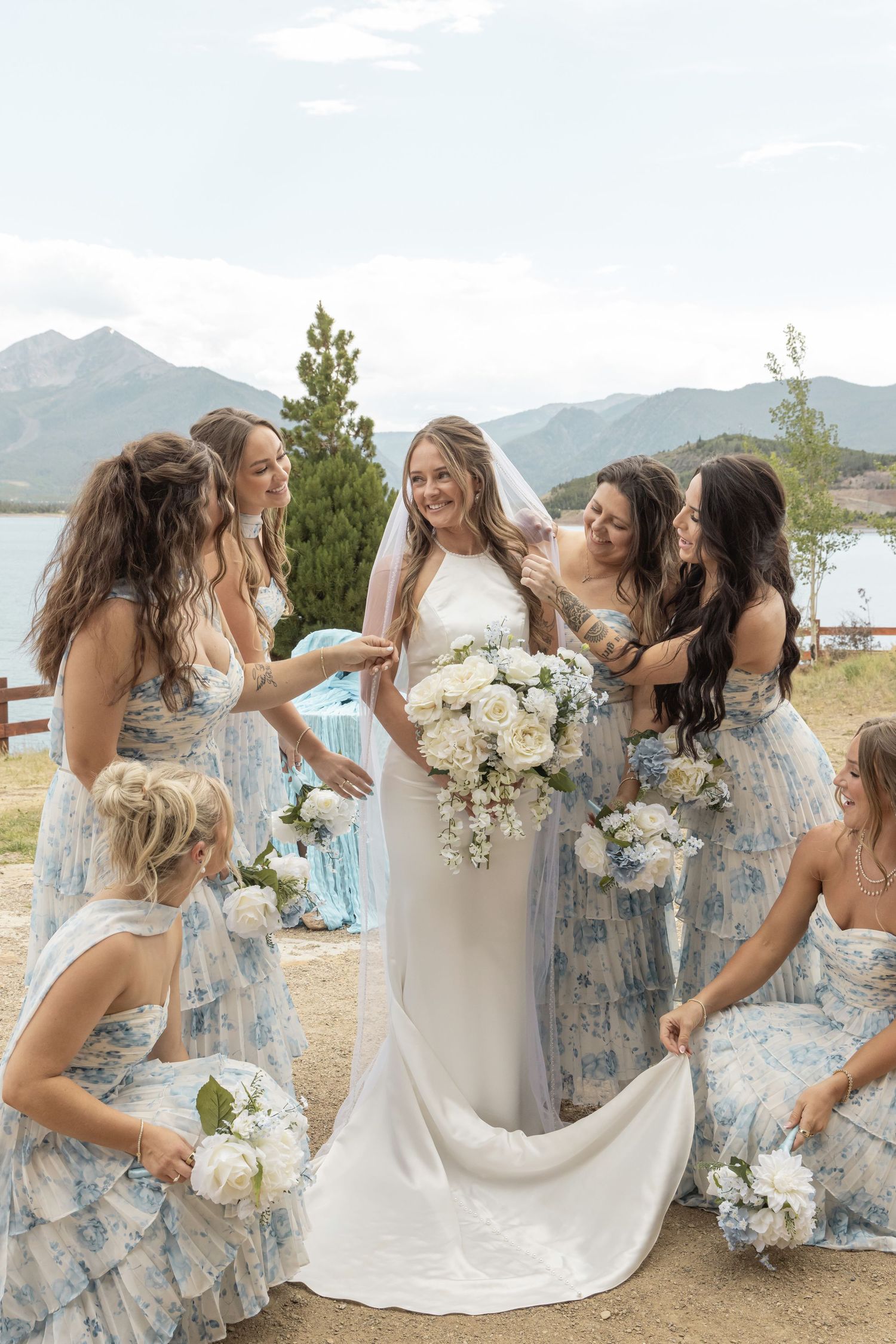 Bride in white gown with bridesmaids in blue floral dresses holding white bouquets near mountain lake backdrop.