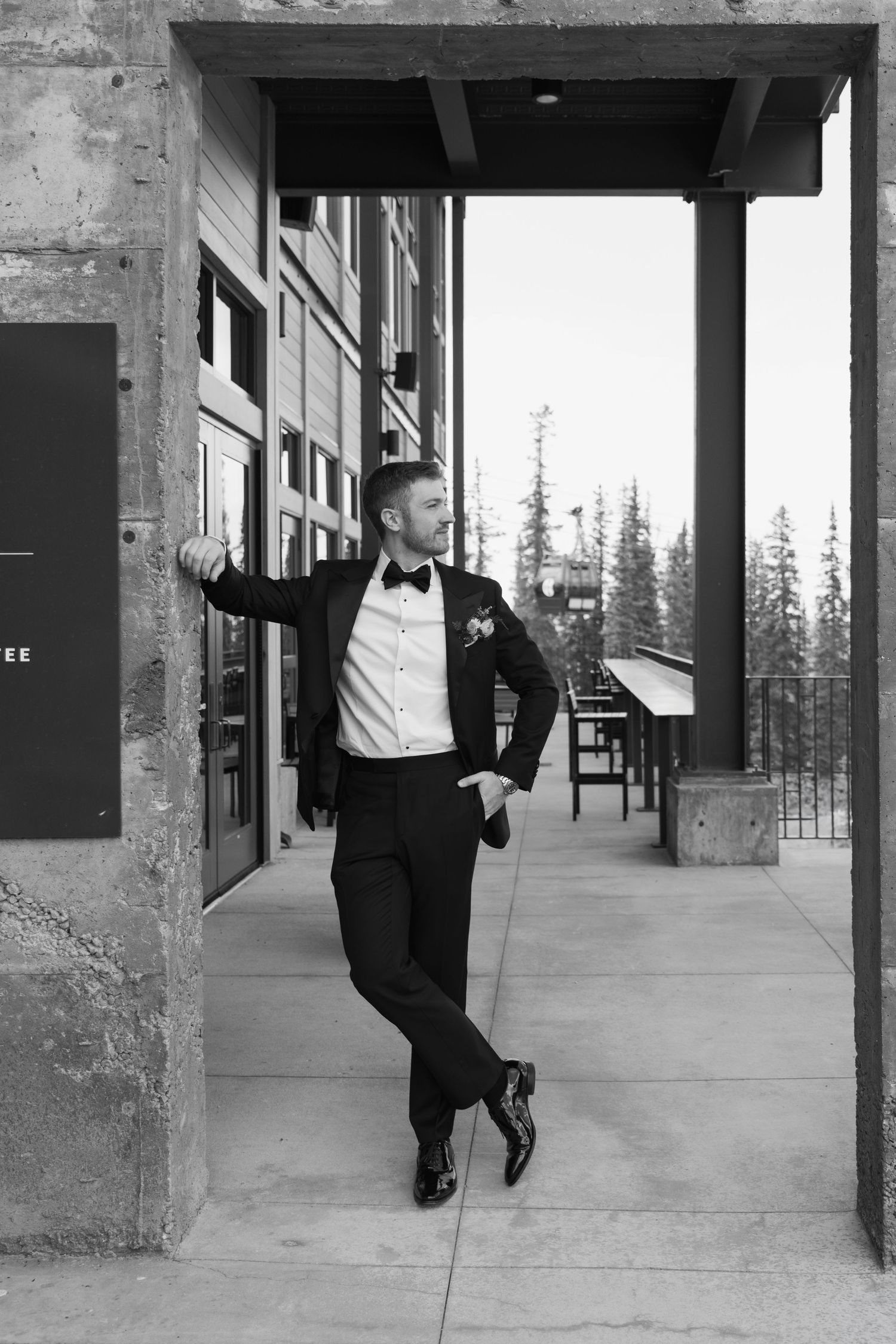 Groom in tuxedo and bow tie poses confidently in a doorway at a mountain wedding venue, black and white photo.