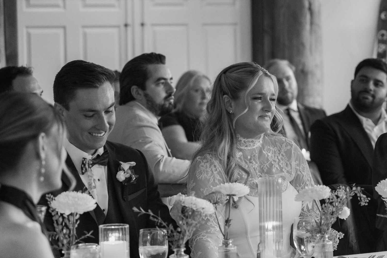 Black and white photo of smiling bride and groom seated at wedding reception table with candles and guests.