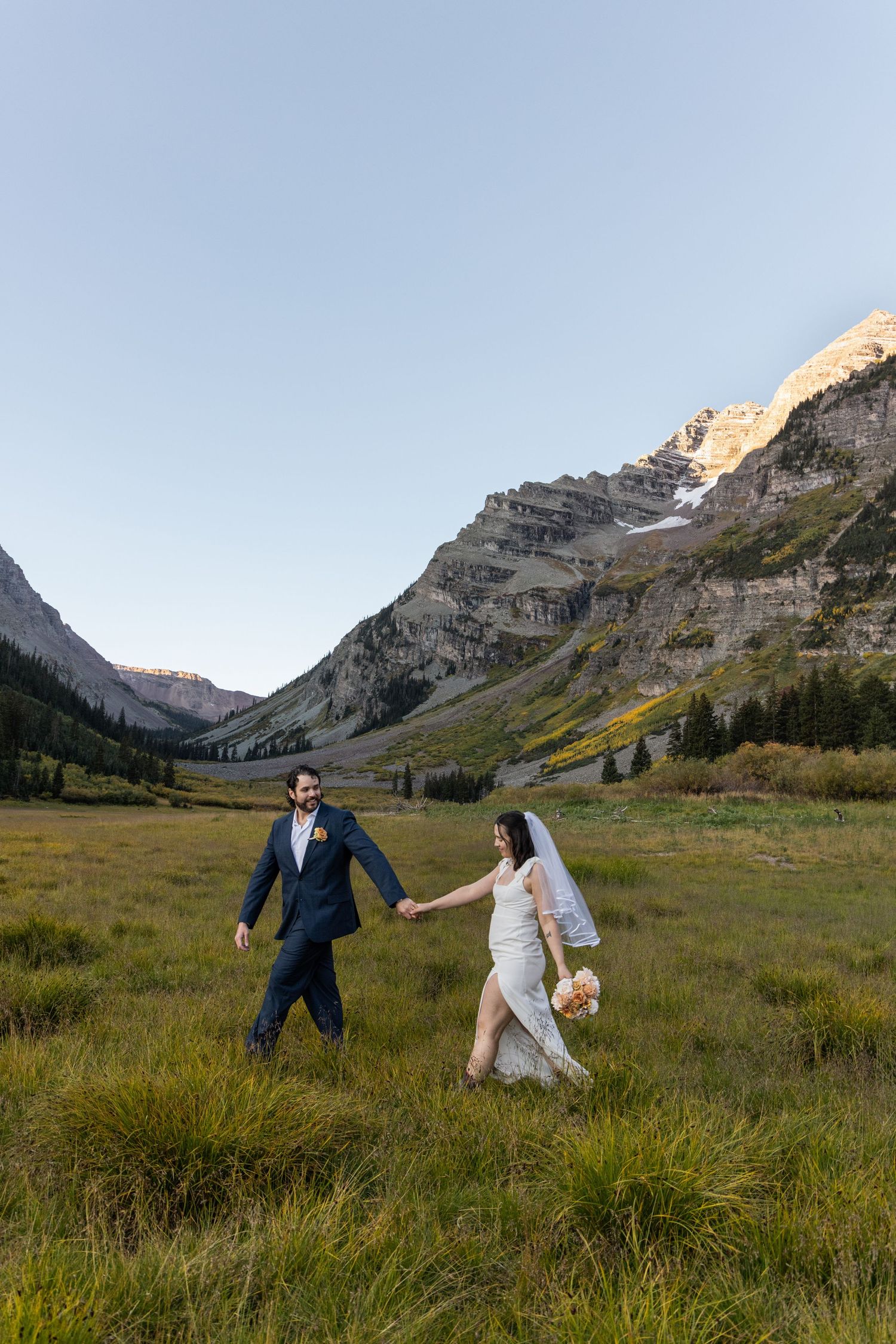 A couple in wedding attire walks through a mountain meadow with dramatic peaks and blue skies in the background.