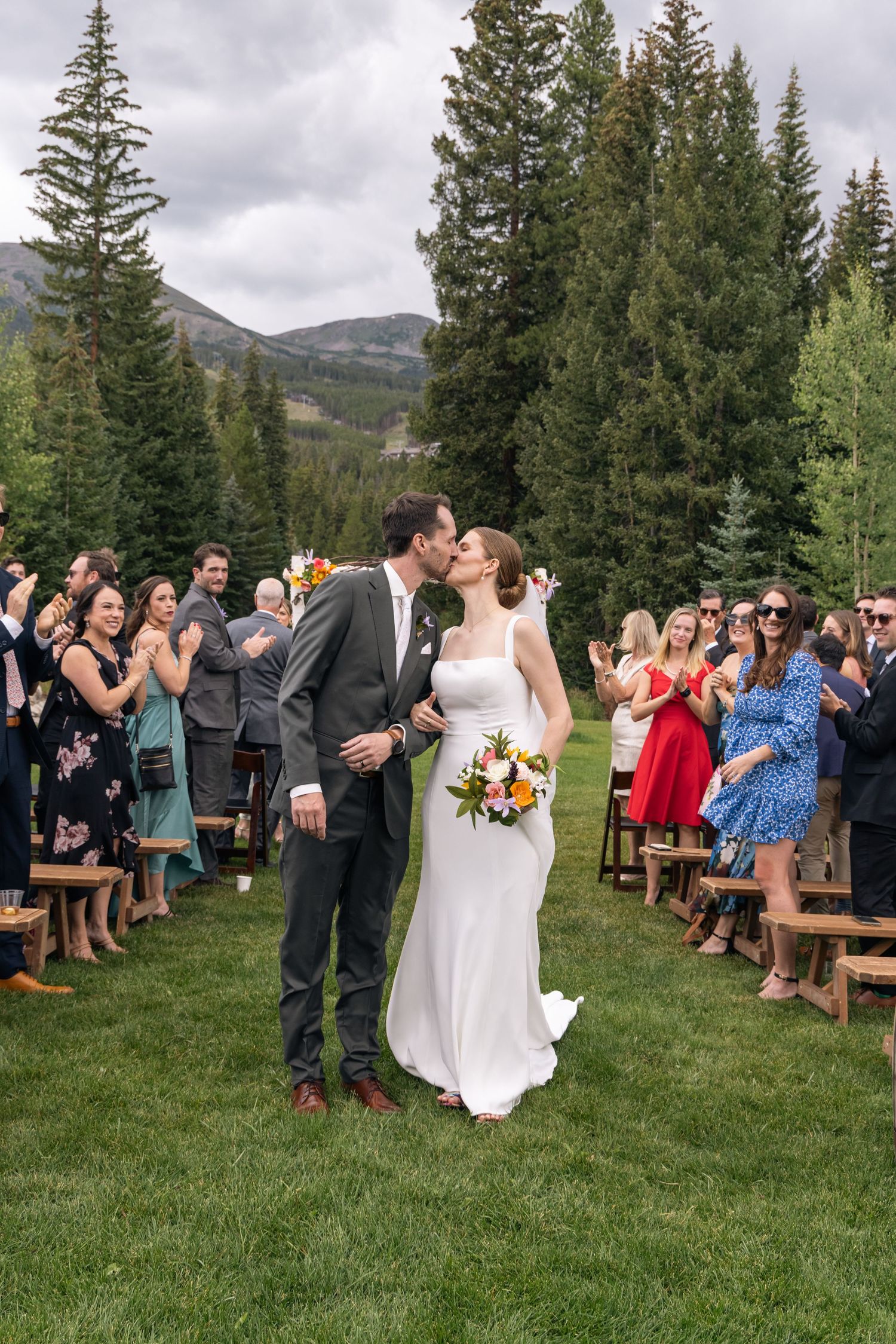Bride and groom share a first kiss as newlyweds walking down the aisle at an outdoor mountain wedding ceremony.