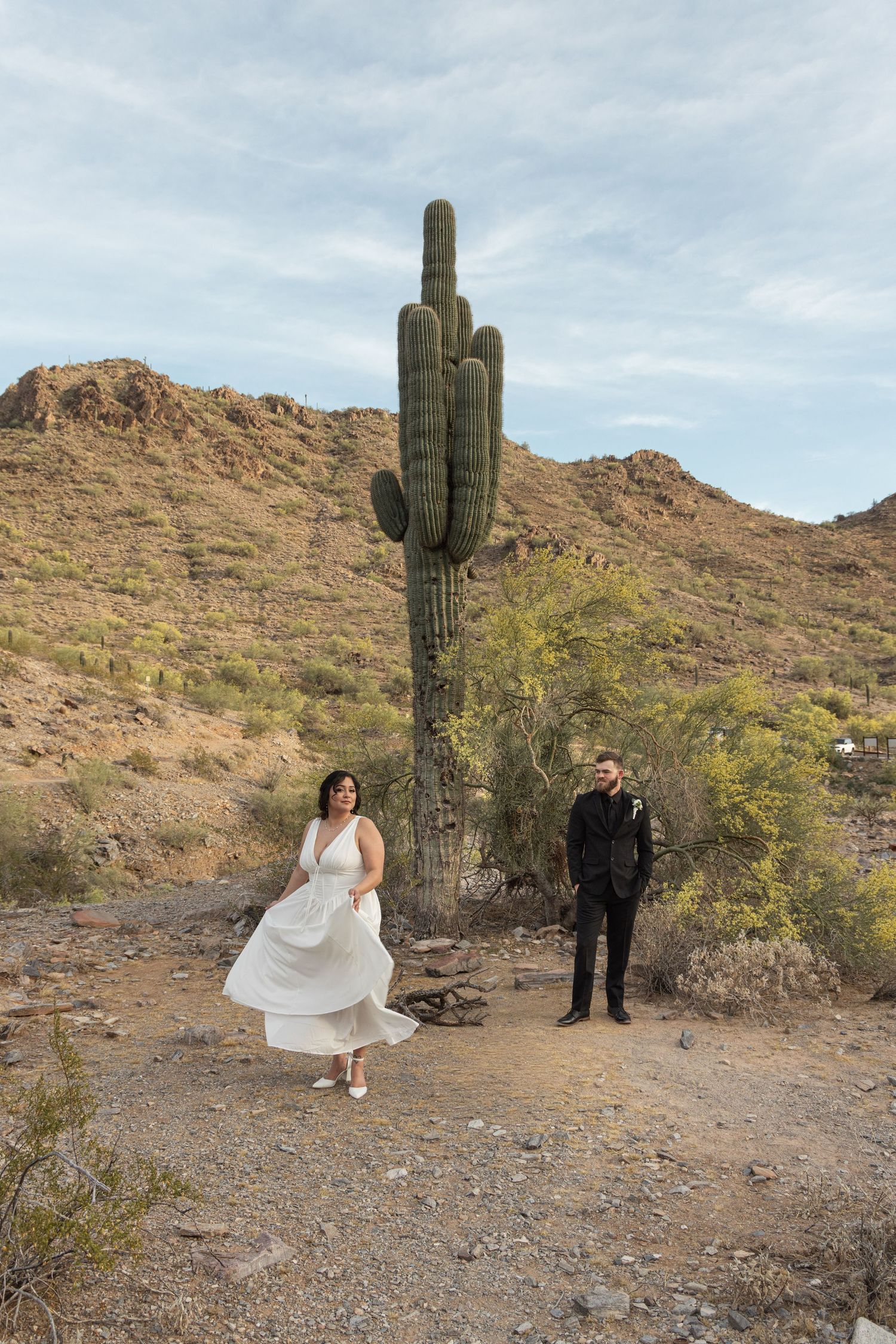 A couple poses for wedding photos in a desert landscape with towering saguaro cactus and rugged mountains in the background.