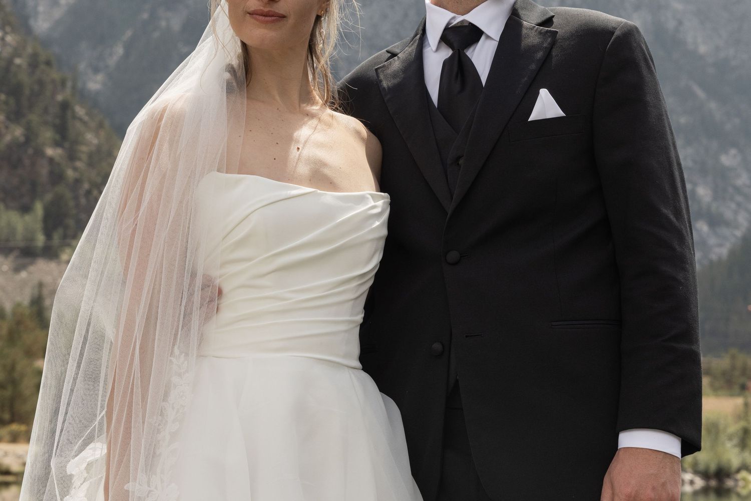 Bride in white strapless gown and veil stands with groom in black tuxedo at mountain wedding ceremony.
