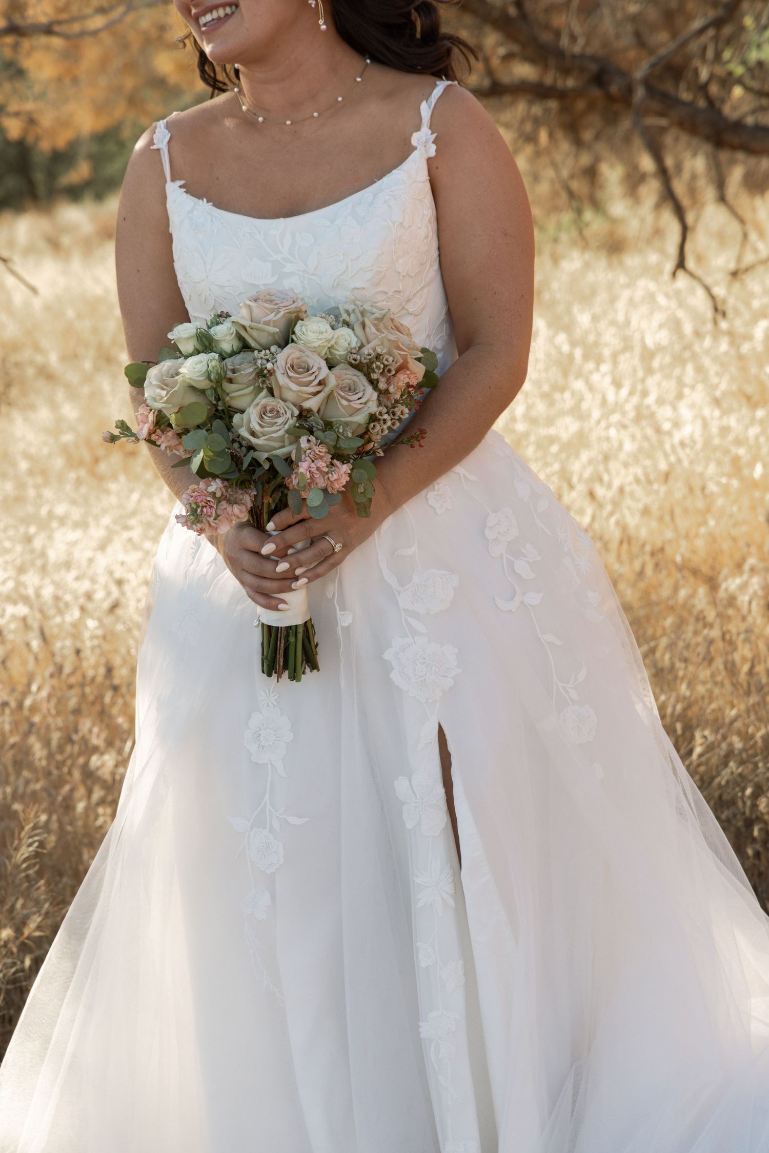 Bride in white lace A-line wedding gown holding blush rose bouquet outdoors in golden field.