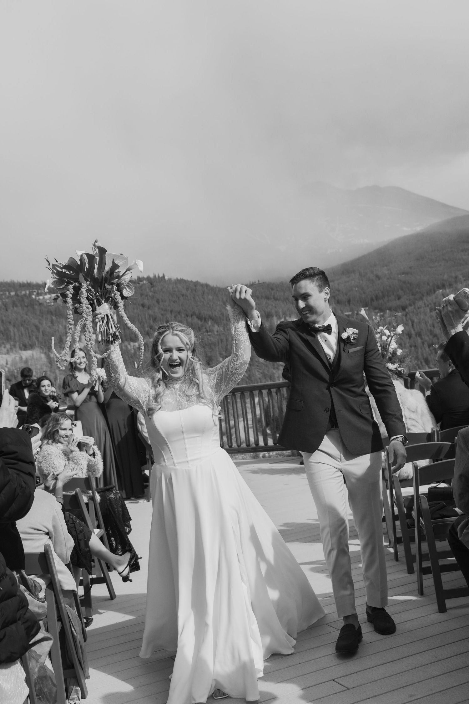 Black and white photo of joyful newlyweds celebrating recessional on mountain venue deck, bride raising bouquet.