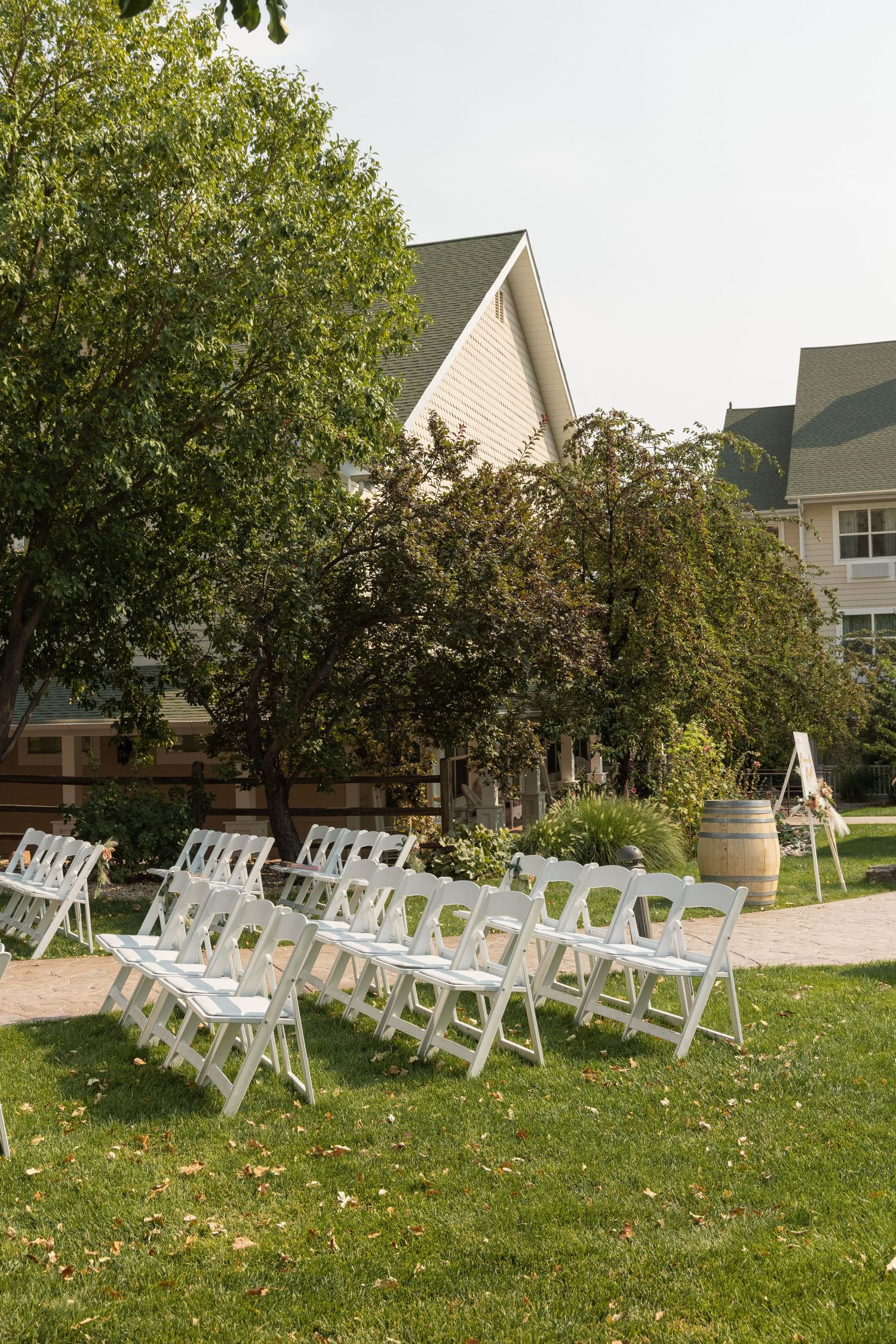 Rows of white folding chairs set up on a green lawn for an outdoor wedding ceremony, with a wine barrel and inn nearby.