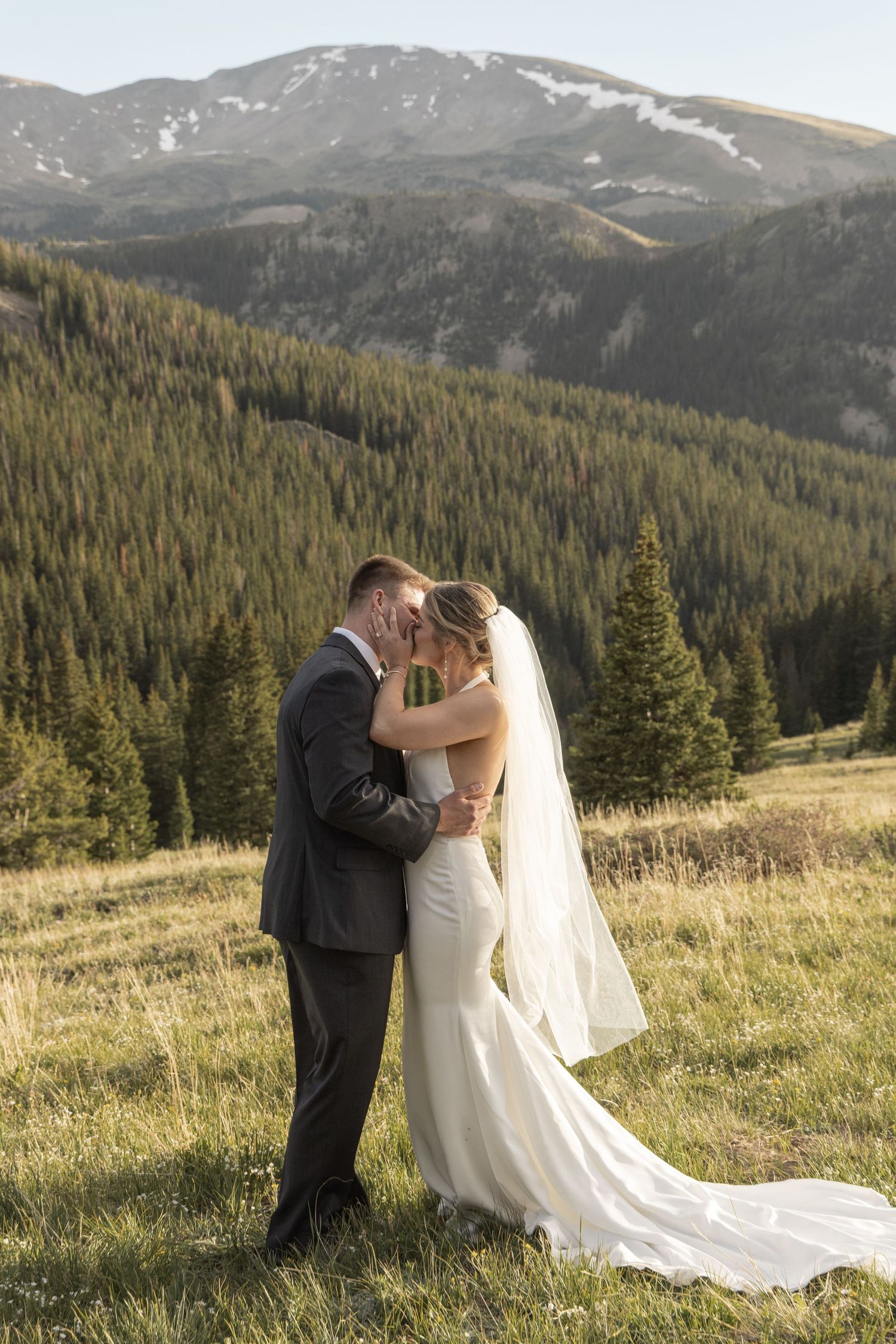 Bride and groom kissing in a mountain meadow, surrounded by evergreen trees and snow-capped peaks.