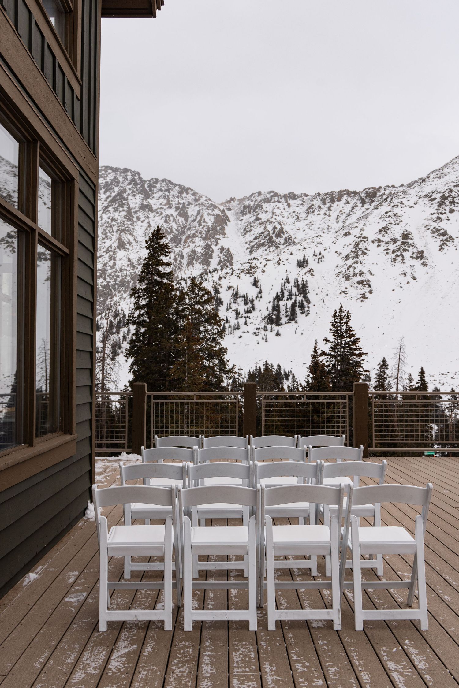 White folding chairs arranged on a snowy mountain deck with evergreen trees and snow-covered peaks in the background.