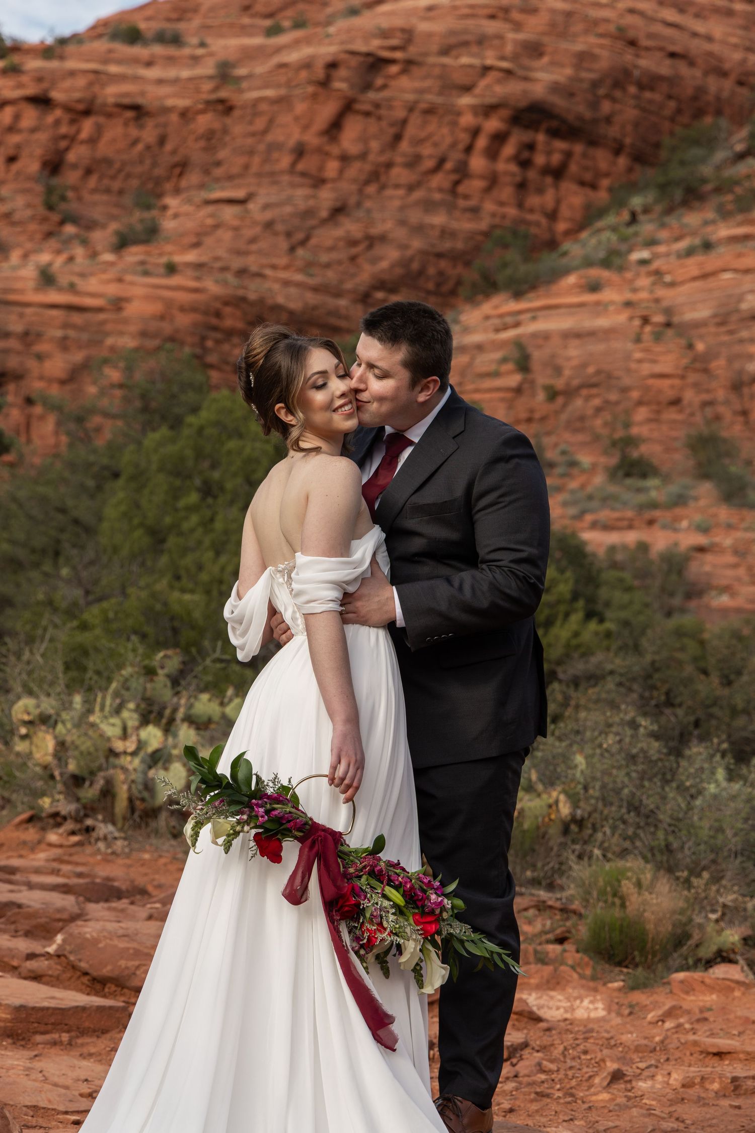 A romantic wedding couple embraces against stunning red rock formations in a desert landscape.