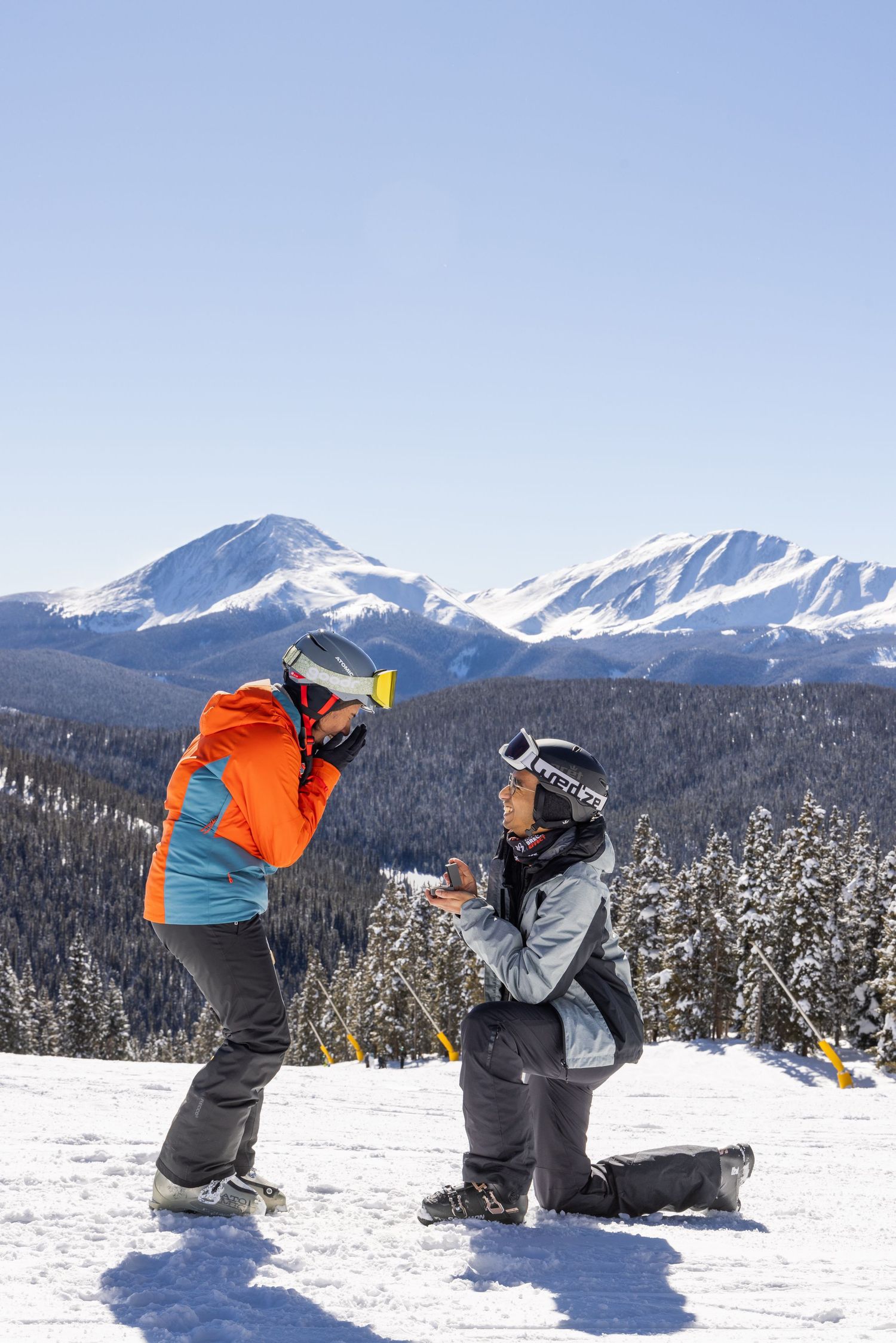 Two snowboarders playfully wrestle in deep powder snow with snow-capped mountains visible in the background.