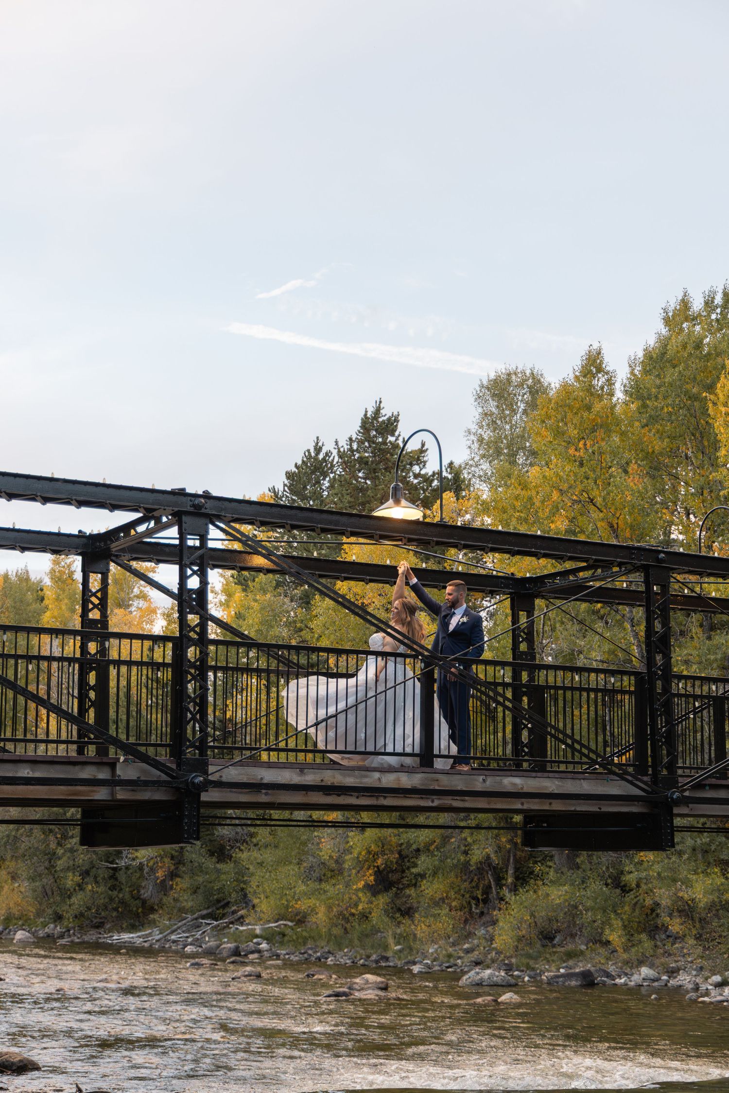 Bride and groom share a romantic moment on a rustic bridge over a river, surrounded by autumn trees.