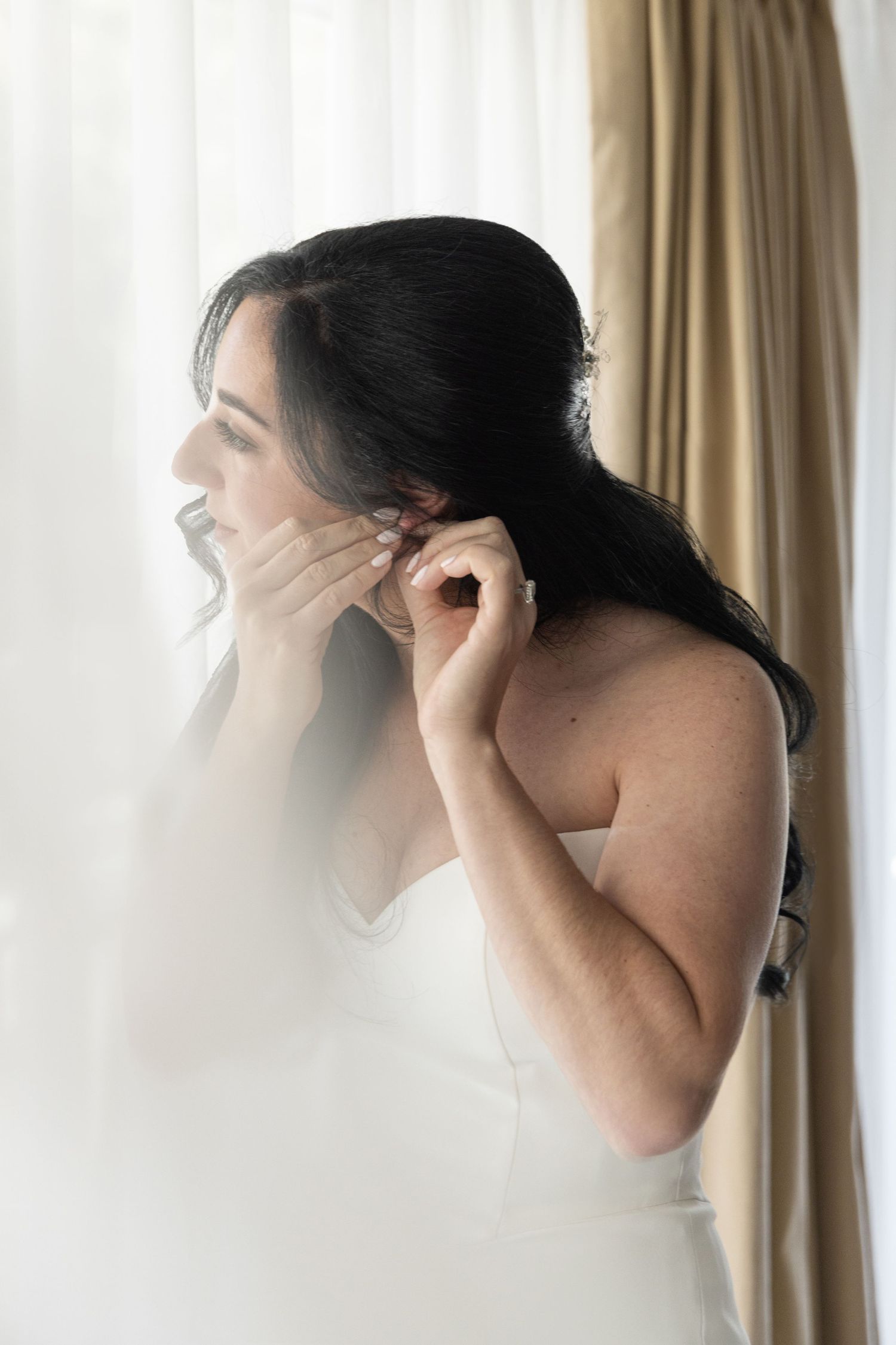 A dark-haired bride in a white dress puts on earrings while standing near a sheer curtained window.