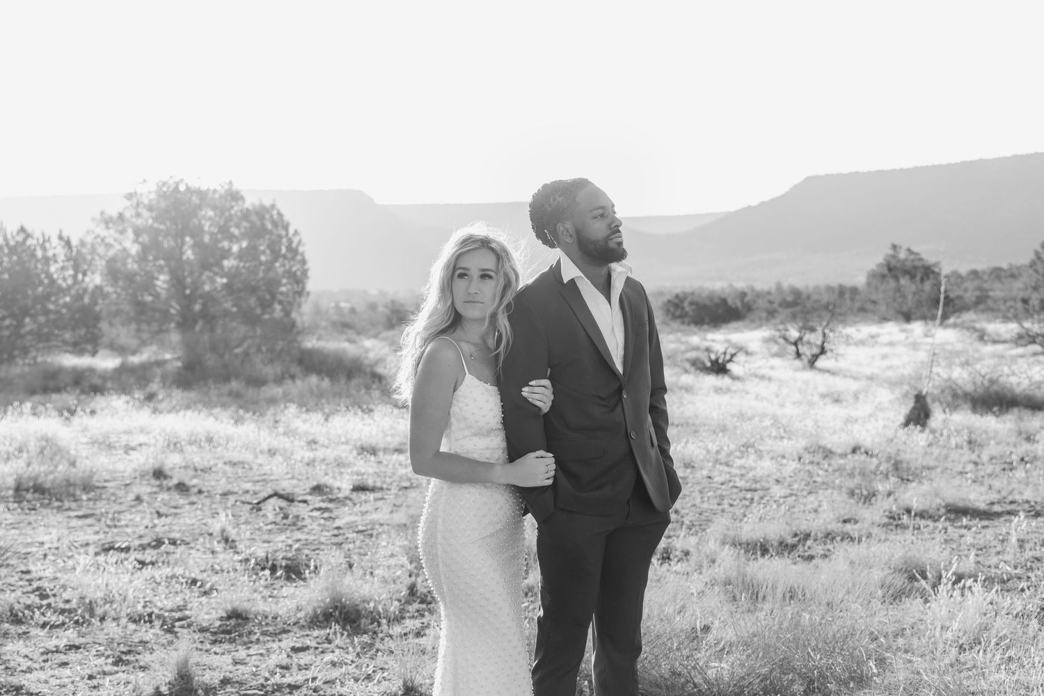 Black and white photo of a couple in wedding attire posing in a desert landscape with mountains in the background.